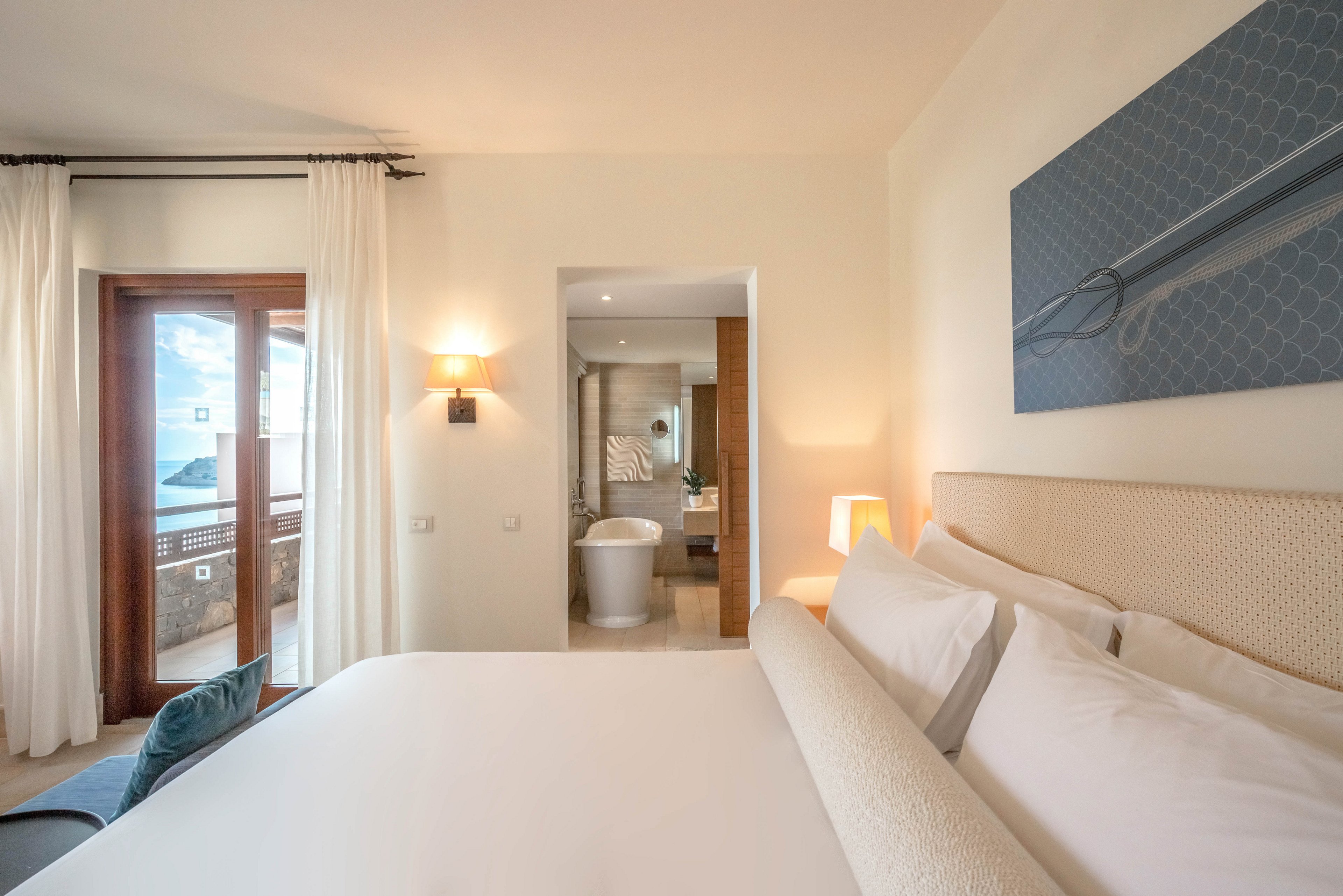 A contemporary hotel bedroom features a large neatly made bed with white linens and a cushioned headboard. Natural light enters through glass doors that open to a balcony with an outdoor view. The room includes warm wall lighting, a bedside lamp, and a blue-toned artwork above the bed. An open doorway reveals an ensuite bathroom with a freestanding bathtub and wooden accents, creating a calm, luxurious atmosphere.