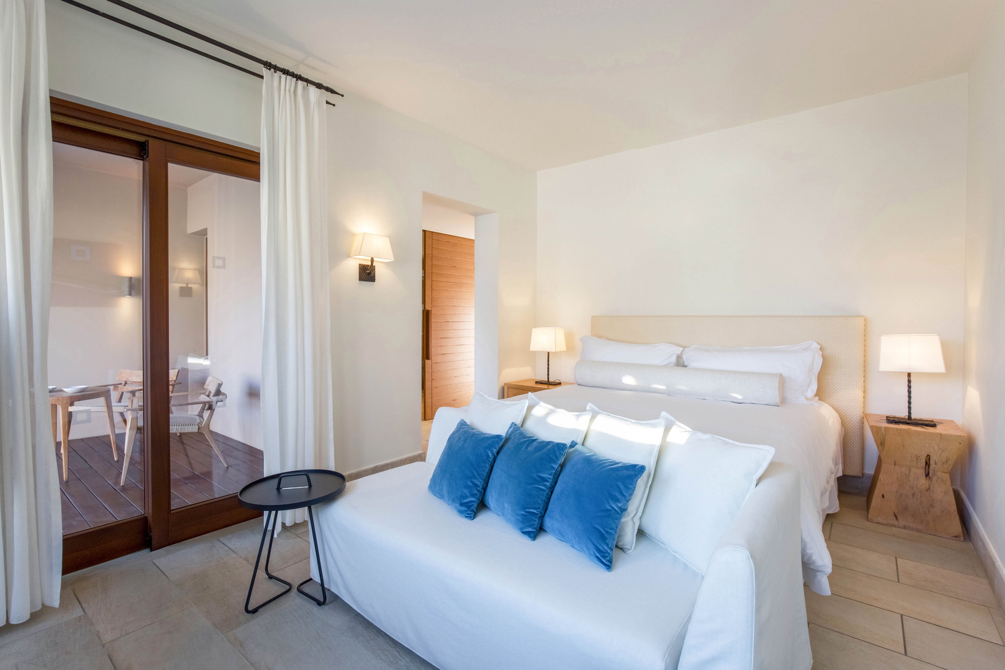 A modern hotel bedroom features a neatly made double bed with white linens and three blue accent pillows. A small white sofa sits at the foot of the bed beside a round black side table. Warm bedside lamps and wall sconces illuminate the neutral, minimalist decor. Large glass doors with white curtains open to an adjacent terrace area with additional seating.