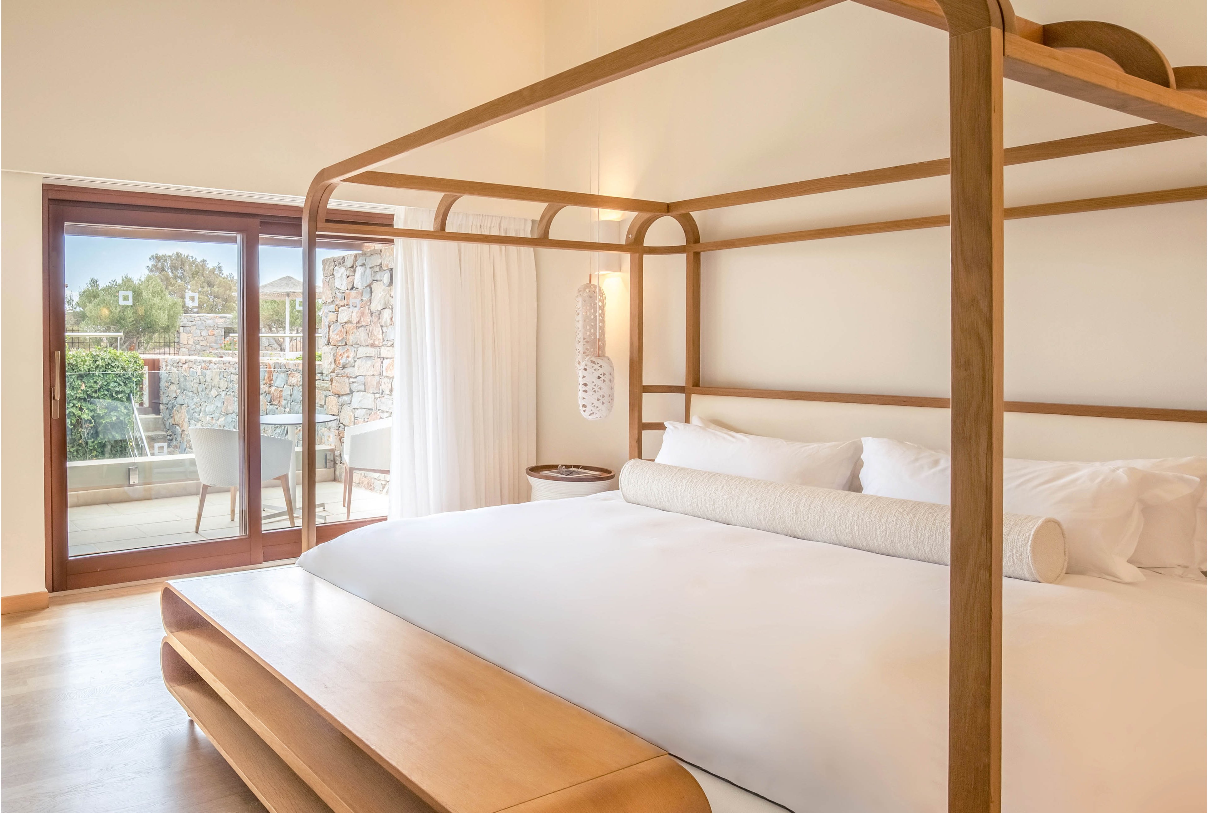 A spacious resort-style bedroom features a large wooden canopy bed with crisp white linens and bolster pillows. Natural light floods the room through wide sliding glass doors that open to a private terrace with outdoor seating. Neutral tones, light wood furniture, and minimal decor create a calm, contemporary atmosphere. A stone wall and greenery are visible outside, enhancing the relaxing vacation setting.