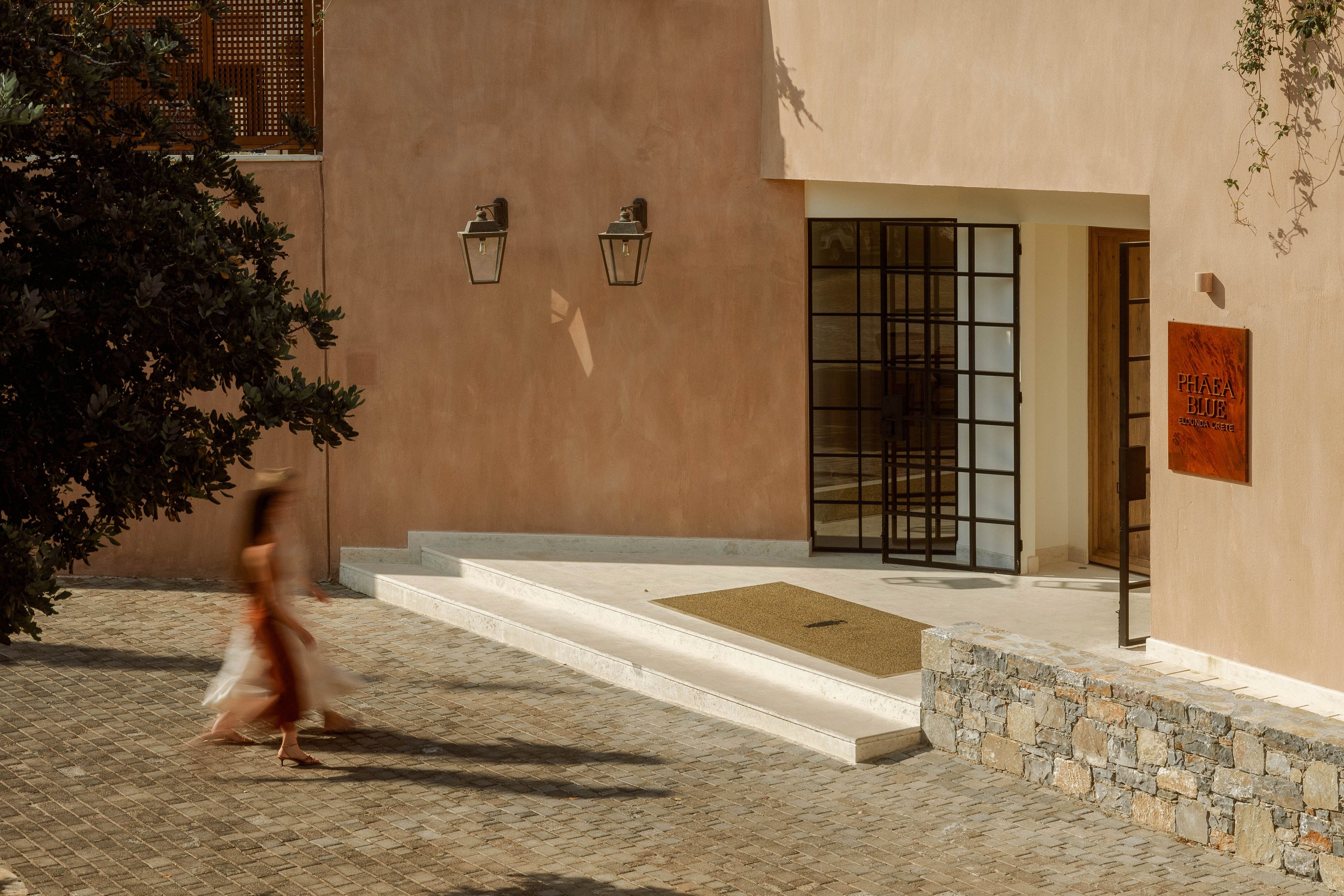 A sunlit Mediterranean-style building with warm terracotta walls stands under a clear sky. The architecture features clean lines, columns, and a stone-paved courtyard. A blurred person in a flowing dress walks across the foreground, adding a sense of motion. Soft shadows, greenery, and distant hills create a calm, vacation-like atmosphere.
