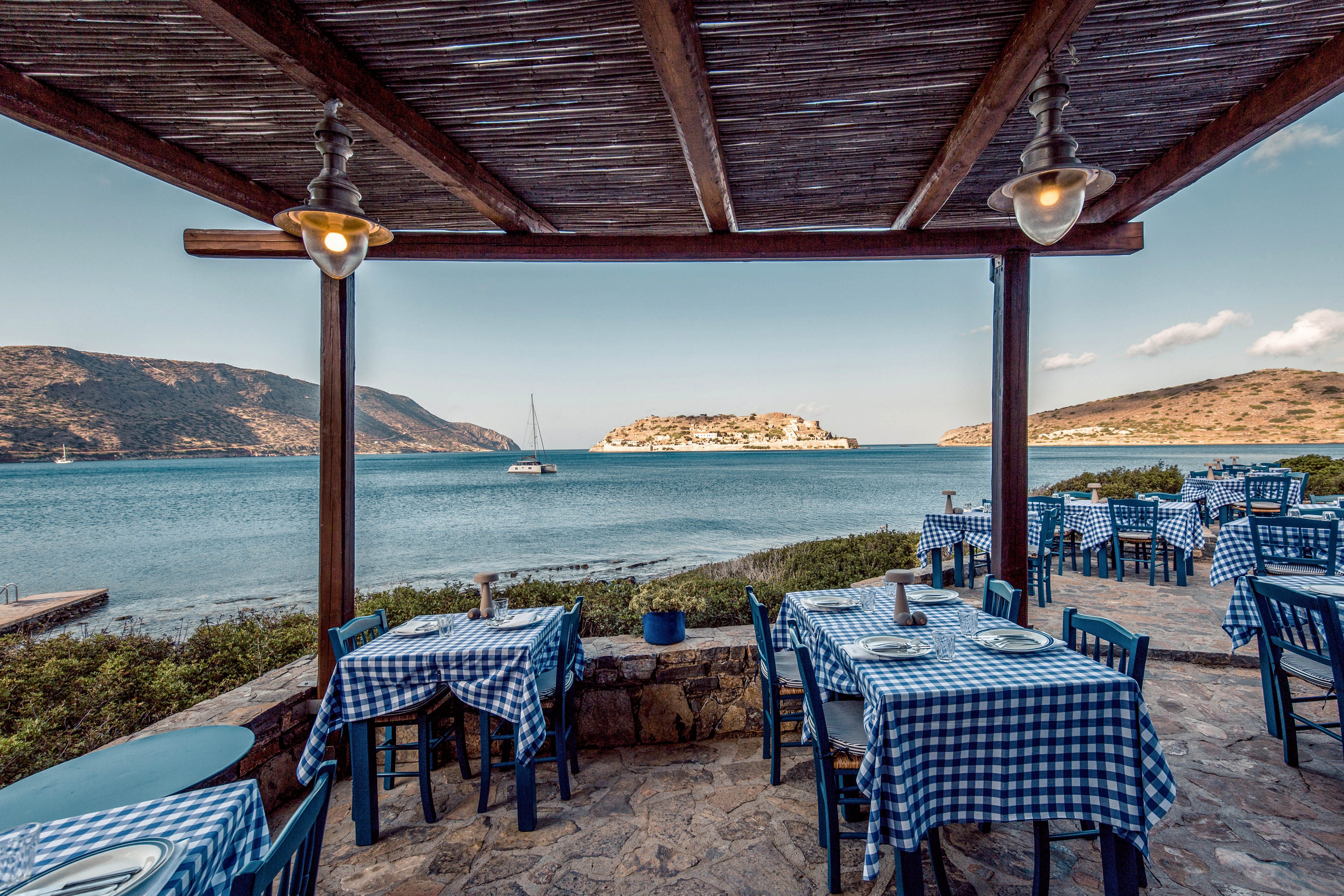 Open-air restaurant tables with blue and white checkered cloths overlook a calm seaside bay. The covered stone terrace frames a view of distant hills and a small island in the background. Empty chairs and neatly set tables suggest a quiet moment before guests arrive. Soft natural light and clear skies create a relaxed Mediterranean holiday atmosphere.