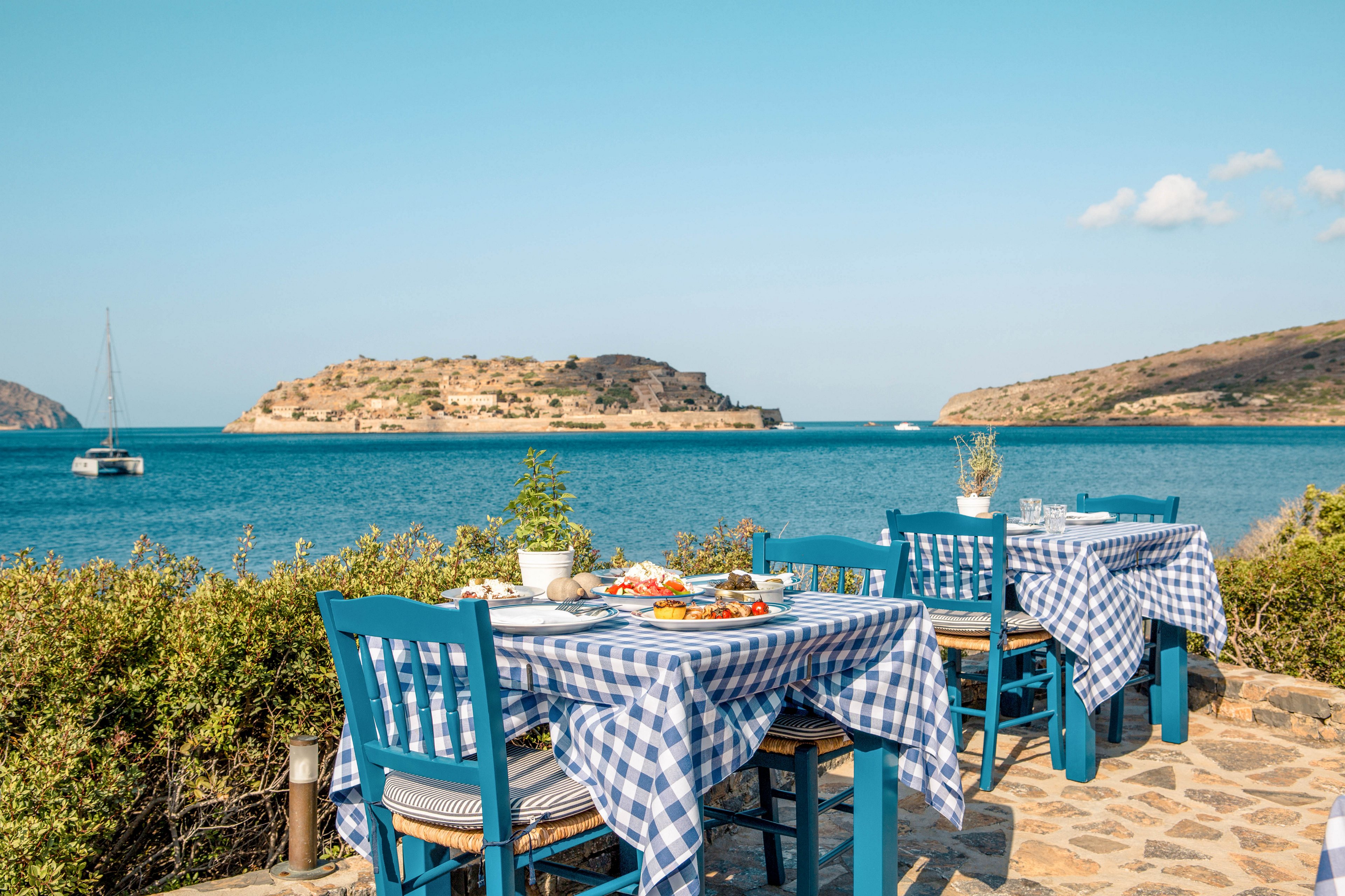 Outdoor restaurant tables with blue chairs overlook a calm turquoise sea and a small island in the distance. The tables are covered with blue and white checkered tablecloths and set with plates of food and drinks. A sailboat floats on the water under a clear blue sky with a few scattered clouds. Low shrubs and stone paving frame the cozy coastal dining area, creating a relaxed Mediterranean atmosphere.