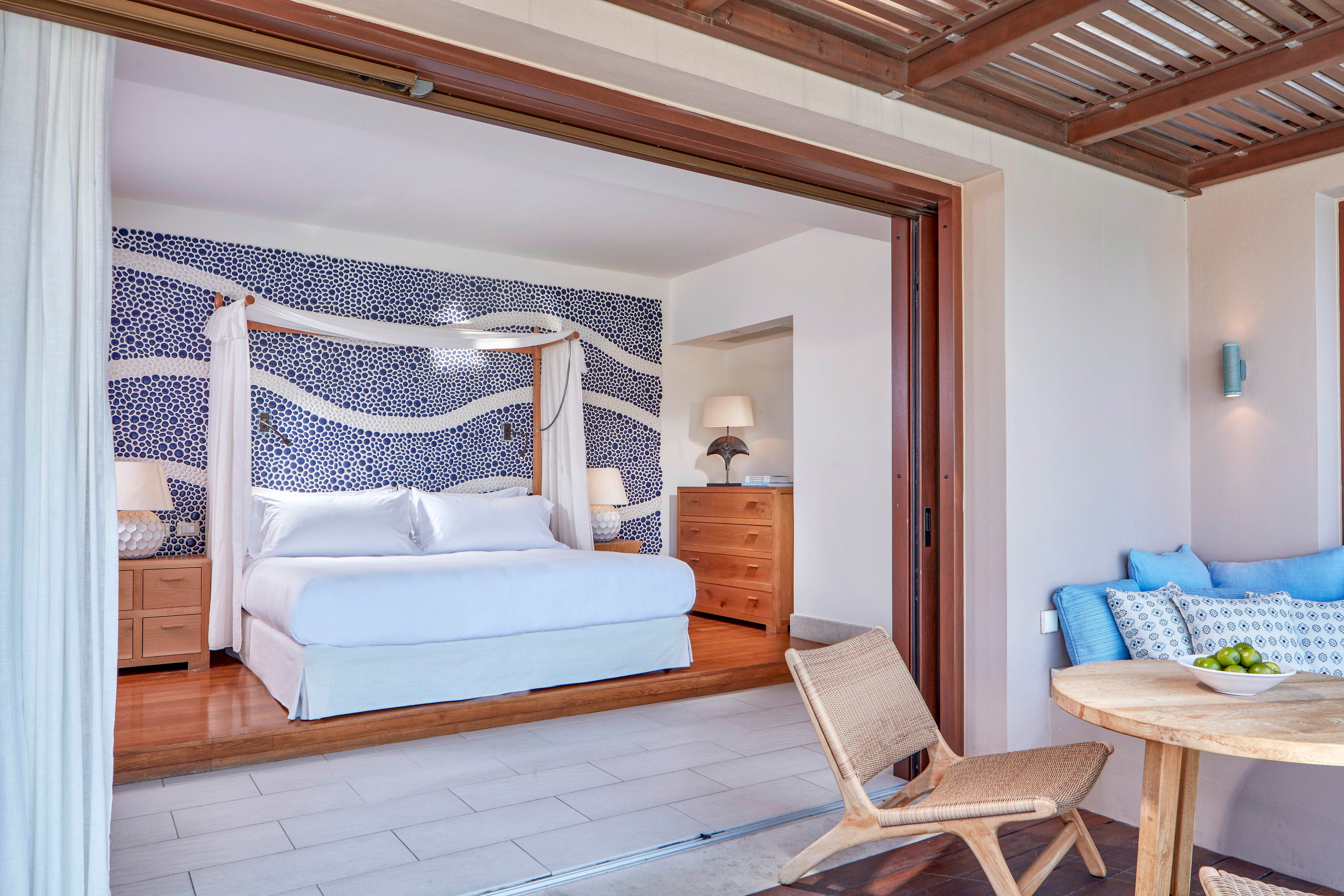 Spacious hotel bedroom featuring a large white bed with a canopy frame is viewed from an open terrace. The interior includes wooden furniture, soft lighting, and a decorative blue and white patterned accent wall. Outside, a small round table with fruit and a cushioned bench create a relaxed sitting area under a wooden pergola. The overall scene feels airy, modern, and inviting, with natural light filling the space.
