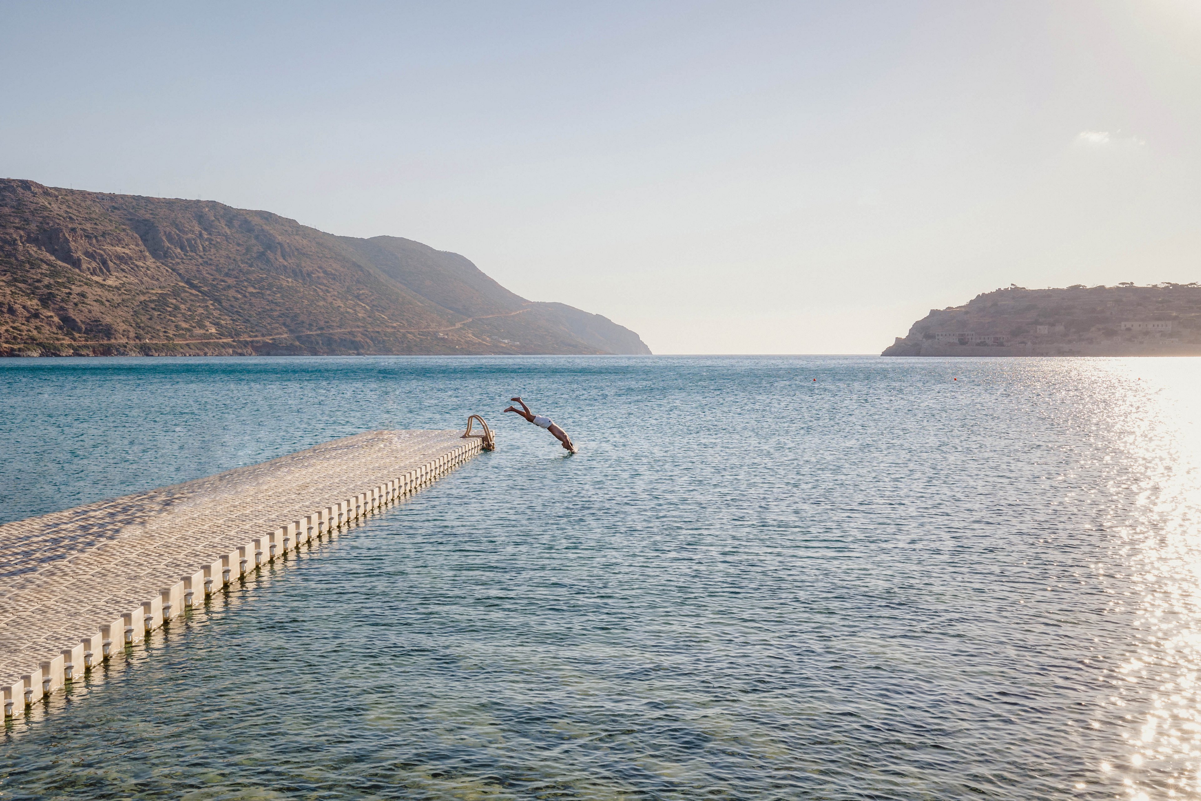 A lone person dives off the end of a narrow pier into a calm, clear sea. The scene is set in a wide coastal bay surrounded by rugged hills and distant shoreline. Gentle ripples on the water and soft sunlight create a peaceful, summery atmosphere. The expansive sky and open water emphasize freedom and relaxation in nature.