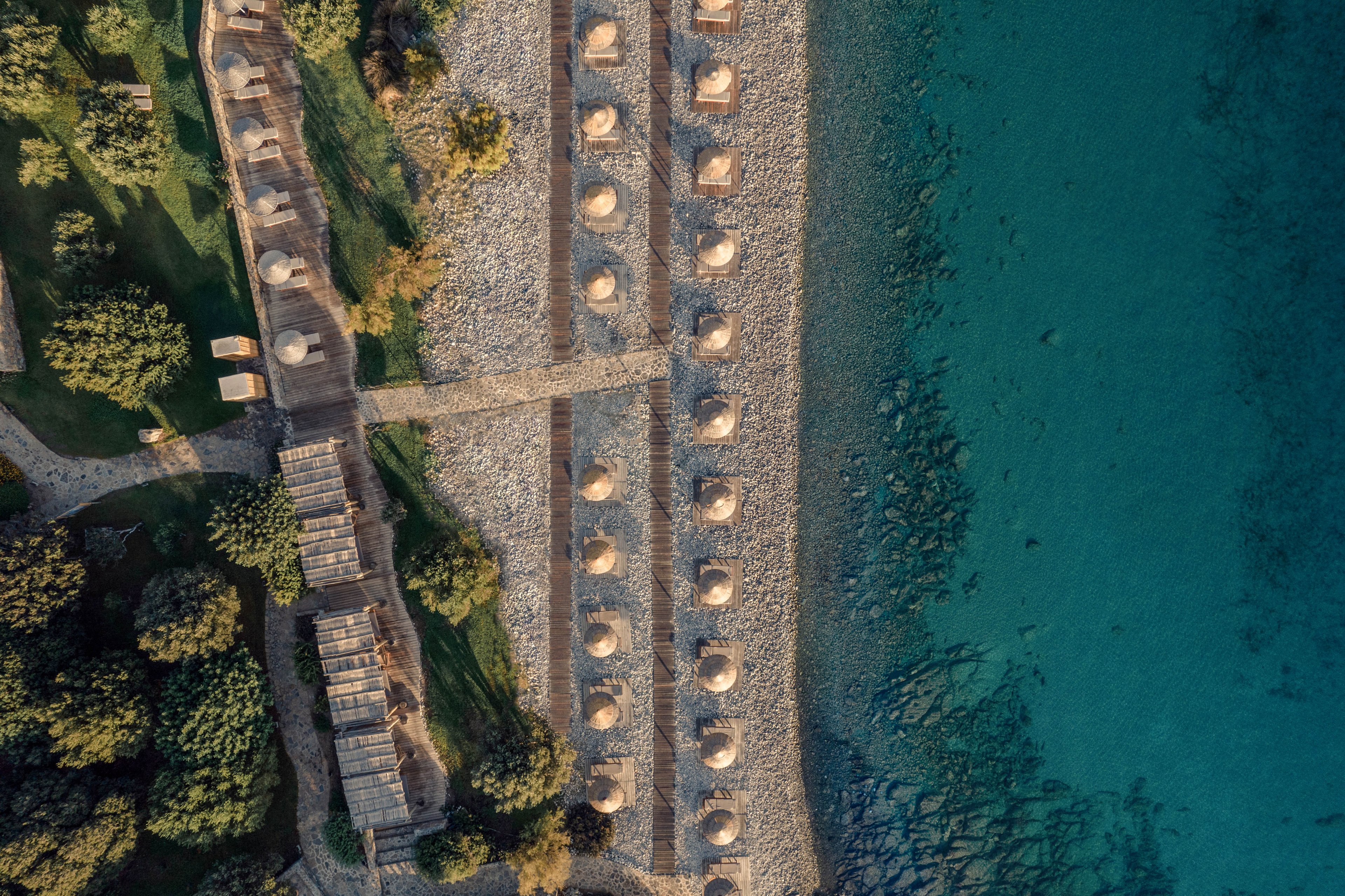 Aerial view of a coastal resort with rows of straw umbrellas neatly arranged on a pebble beach. A stone walkway and wooden structures separate the beach area from lush green trees and landscaped grounds. The clear turquoise water reveals the rocky shoreline beneath the surface. The image emphasizes symmetry, natural textures, and the contrast between land and sea.