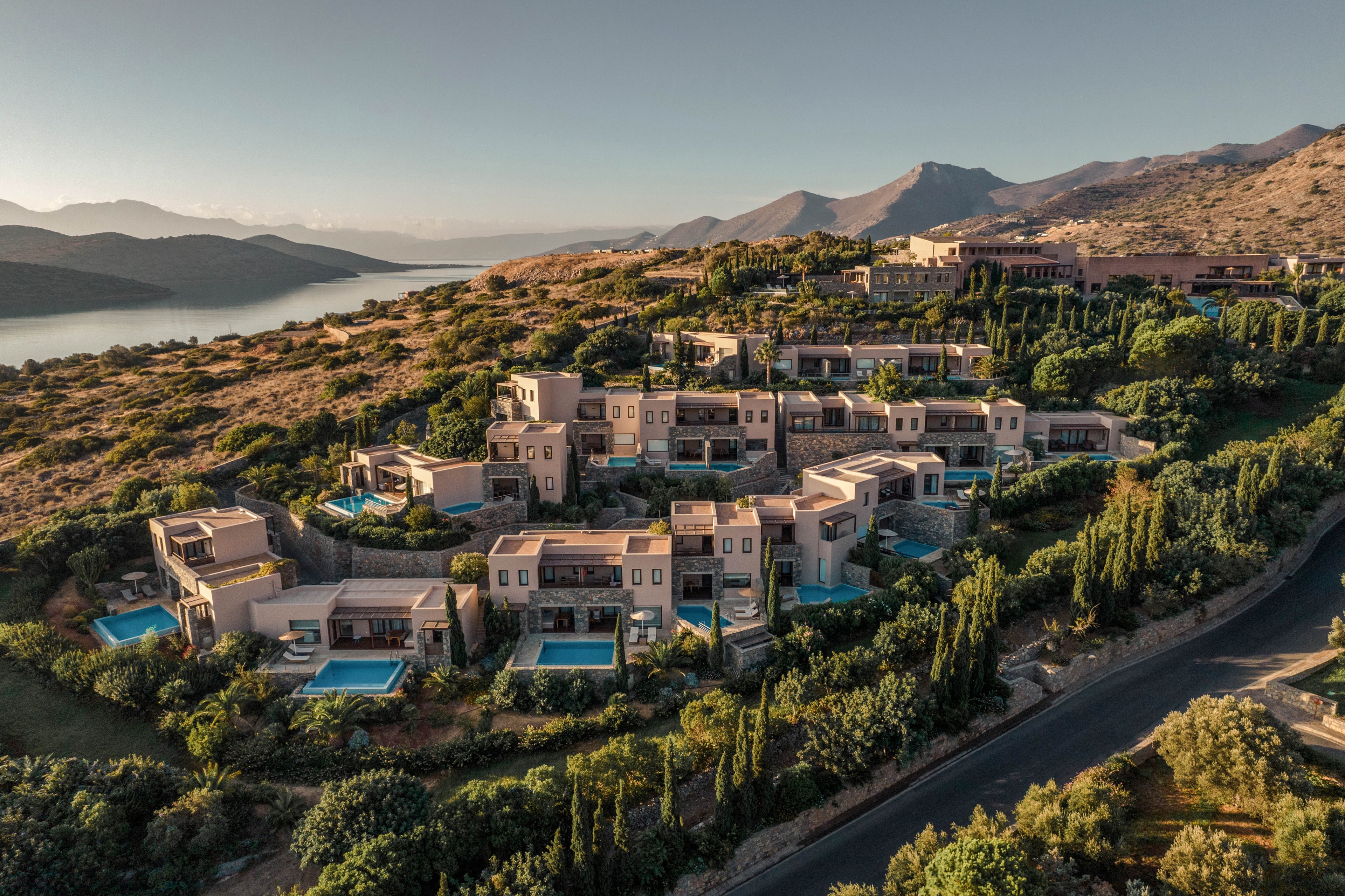A cluster of modern hillside villas with private pools is nestled among trees and shrubs on a rugged slope. The resort-style complex overlooks a calm coastal bay with mountains in the distance. Warm evening light casts long shadows across the buildings and surrounding landscape. The scene conveys a tranquil, upscale vacation destination in a Mediterranean-like setting.
