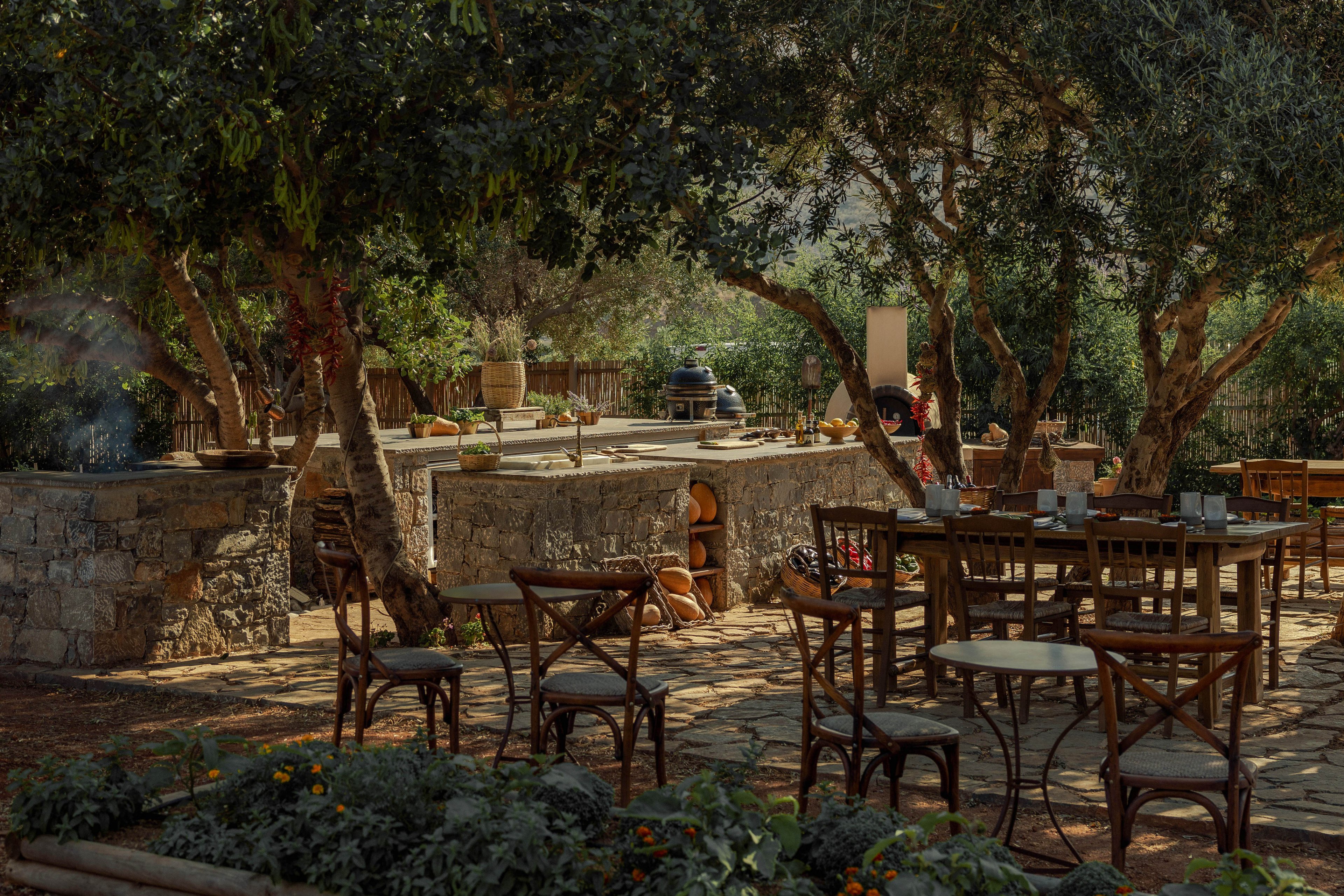 An outdoor café is set beneath tall leafy trees, with wooden tables and chairs arranged on a sunlit patio. A stone wall and counter area create a rustic backdrop, suggesting a relaxed dining or coffee spot. Flower beds and greenery border the seating area, adding color and a natural feel. The warm daylight and dappled shade evoke a calm, leisurely atmosphere suitable for socializing or quiet relaxation.