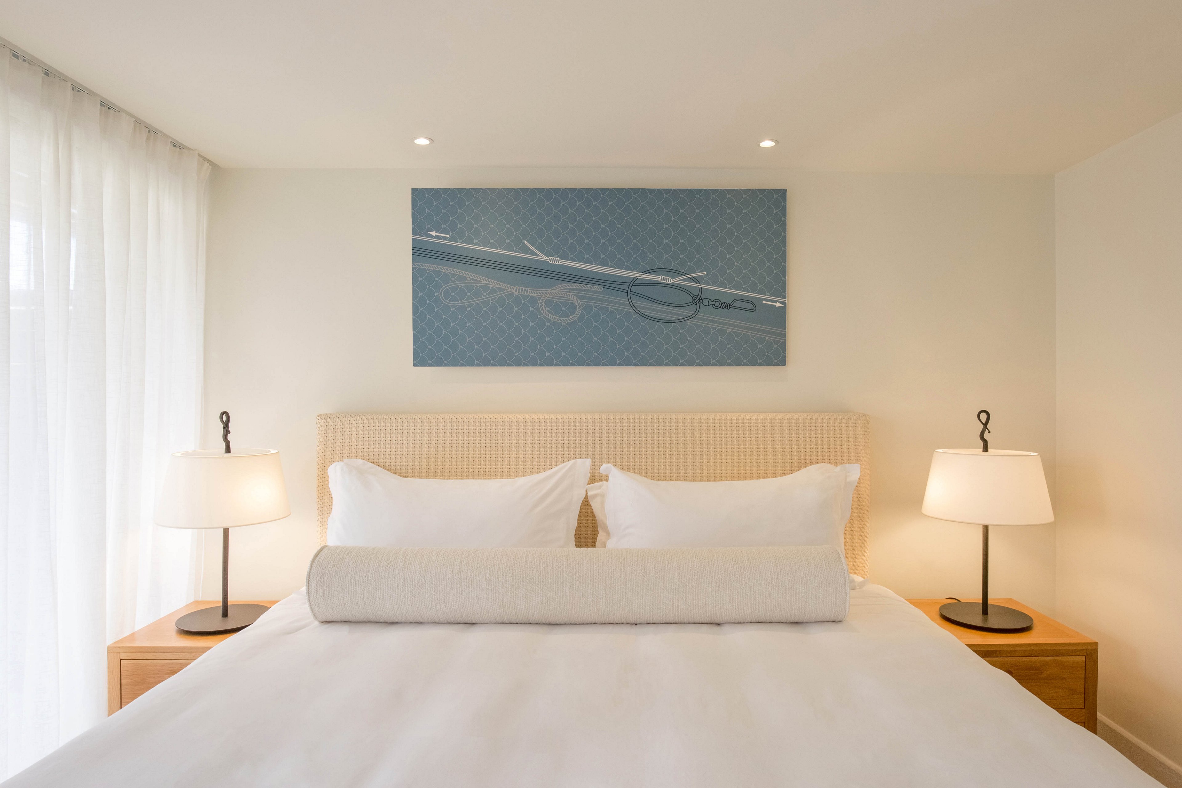 A neatly made bed with crisp white linens and a cylindrical bolster pillow is centered in a minimalist bedroom. Two matching bedside tables hold modern lamps that cast a warm, soft light. Sheer curtains allow natural daylight to filter into the room from the left side. A contemporary artwork featuring a stylized object hangs above the headboard, completing the clean and serene decor.