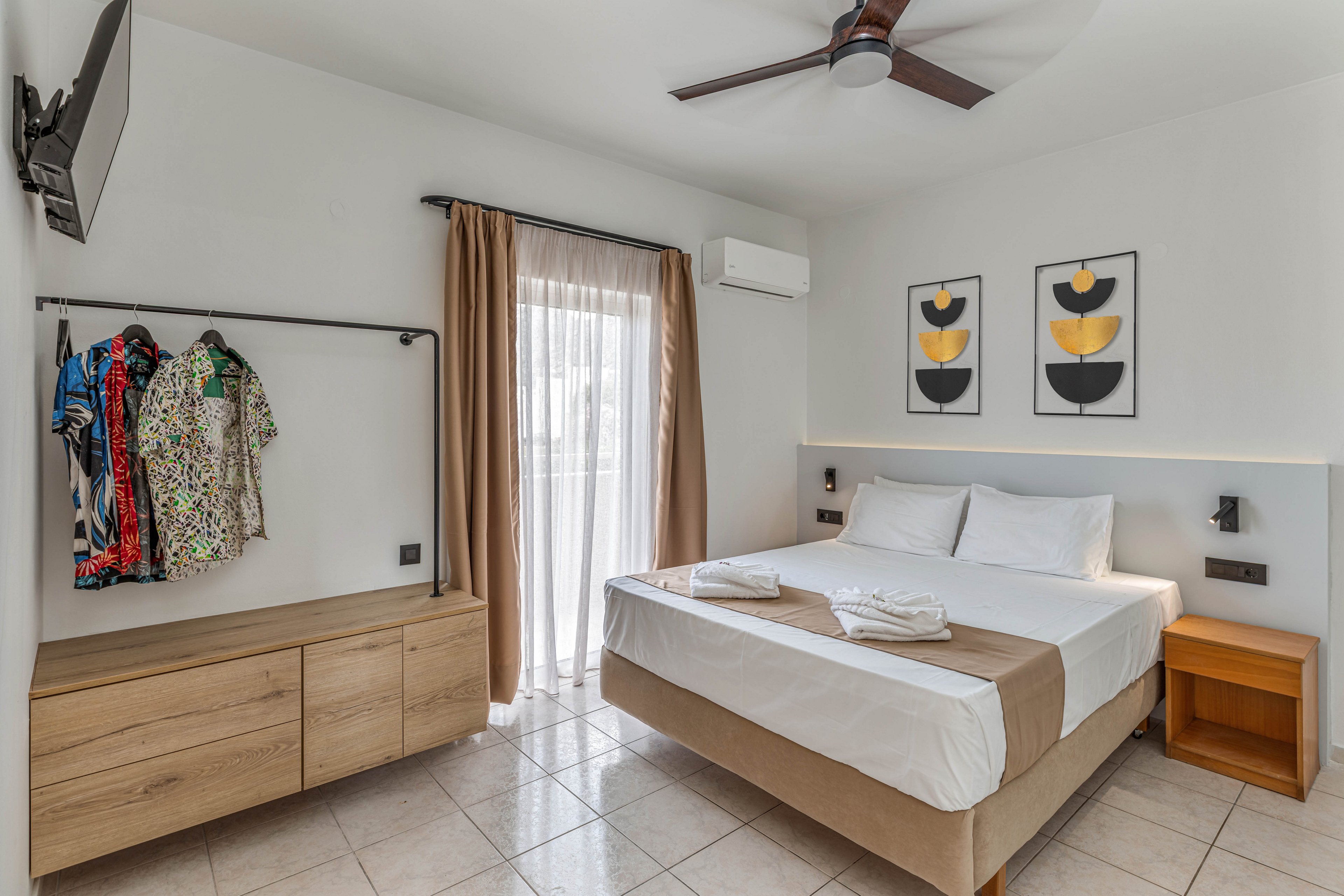 A bright, modern bedroom features a neatly made double bed with white linens and a beige runner. The room includes a wooden dresser, open clothes rack with colorful shirts, and a small bedside table. Natural light enters through a glass door with sheer and brown curtains leading to a balcony. Minimalist artwork, a ceiling fan, wall-mounted TV, and air conditioning unit complete the clean, contemporary look.