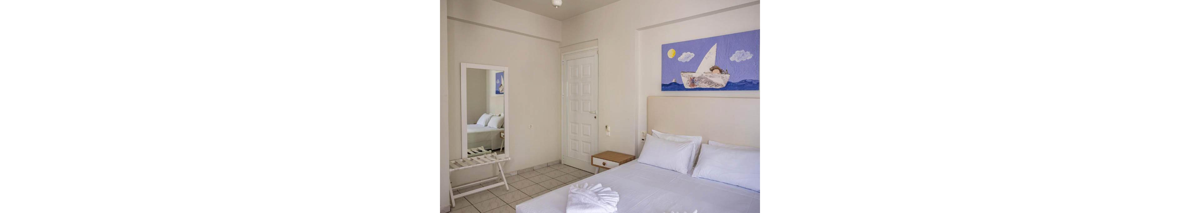 A small, bright bedroom features a neatly made double bed with white linens and pillows. The room has light-colored walls, tiled flooring, and a simple bedside table next to the bed. A full-length mirror reflects part of the bed and a white luggage rack beneath it. Above the headboard hangs a playful nautical painting of a child in a sailboat under a yellow sun and blue sky, adding color to the otherwise neutral space.