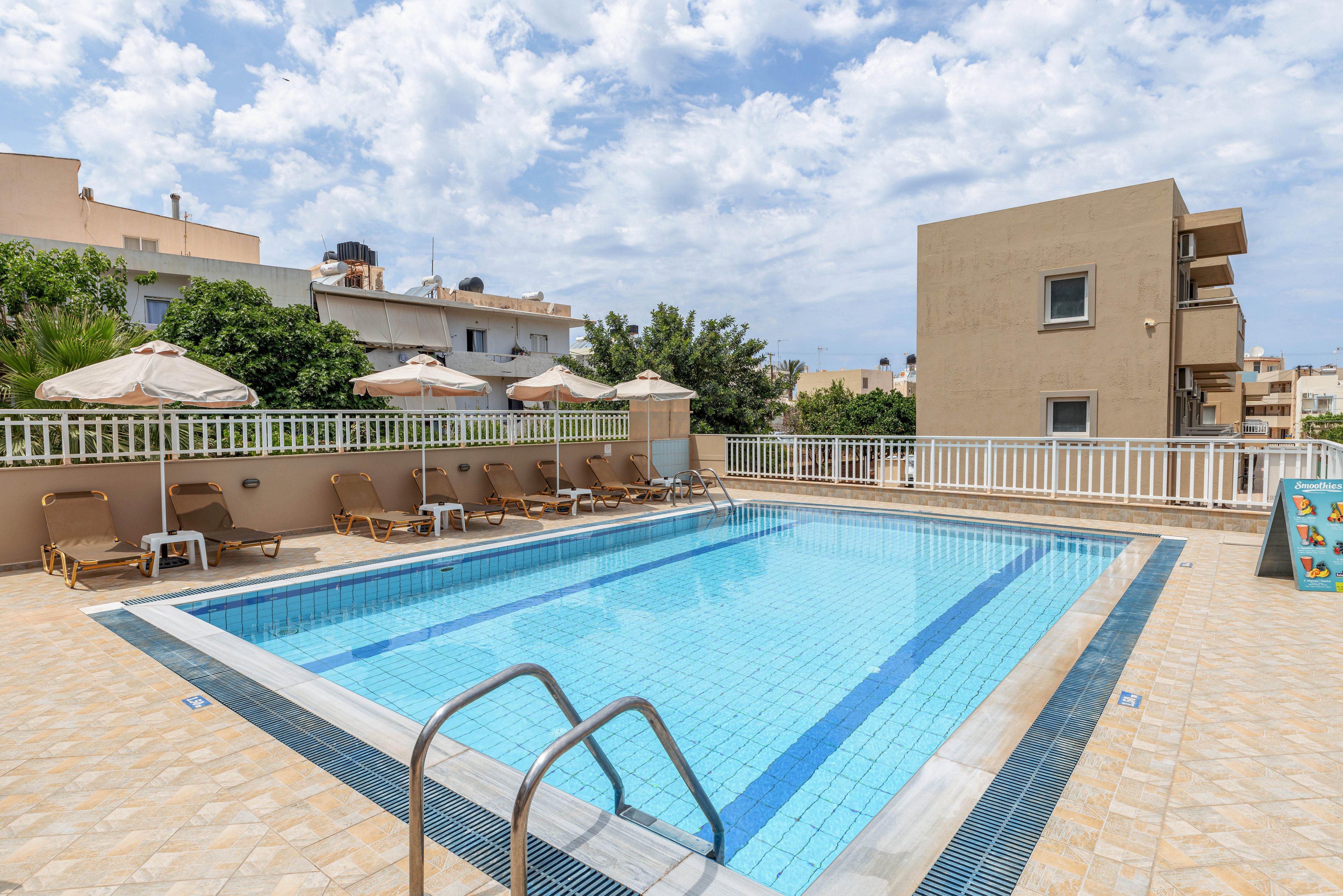 An outdoor rectangular swimming pool is surrounded by a tiled deck and metal safety railings. Several sun loungers with beige umbrellas line one side of the pool, suggesting a hotel or apartment complex setting. Low-rise residential buildings and greenery appear in the background under a partly cloudy sky. The clear blue water and light tiles create a bright, inviting atmosphere for relaxation.