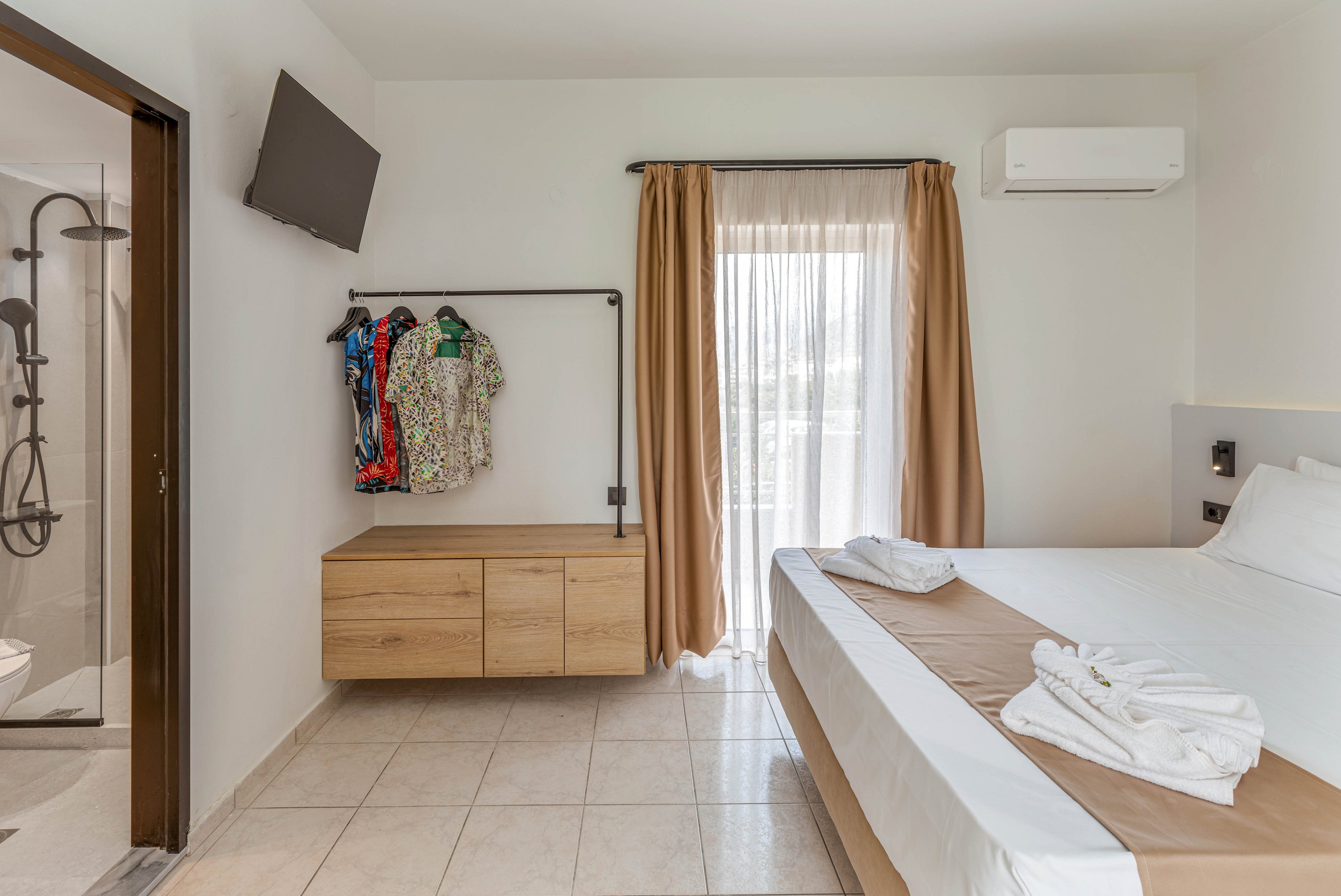 A bright modern hotel bedroom features a large neatly made bed with folded white towels on top. To the side, a wall-mounted wooden cabinet and open clothes rail display several colorful shirts. Floor-to-ceiling curtains cover a glass door leading to a balcony, allowing natural light to fill the room. A wall-mounted television and air conditioning unit emphasize comfort and convenience, while a doorway reveals a contemporary bathroom with a glass shower enclosure.