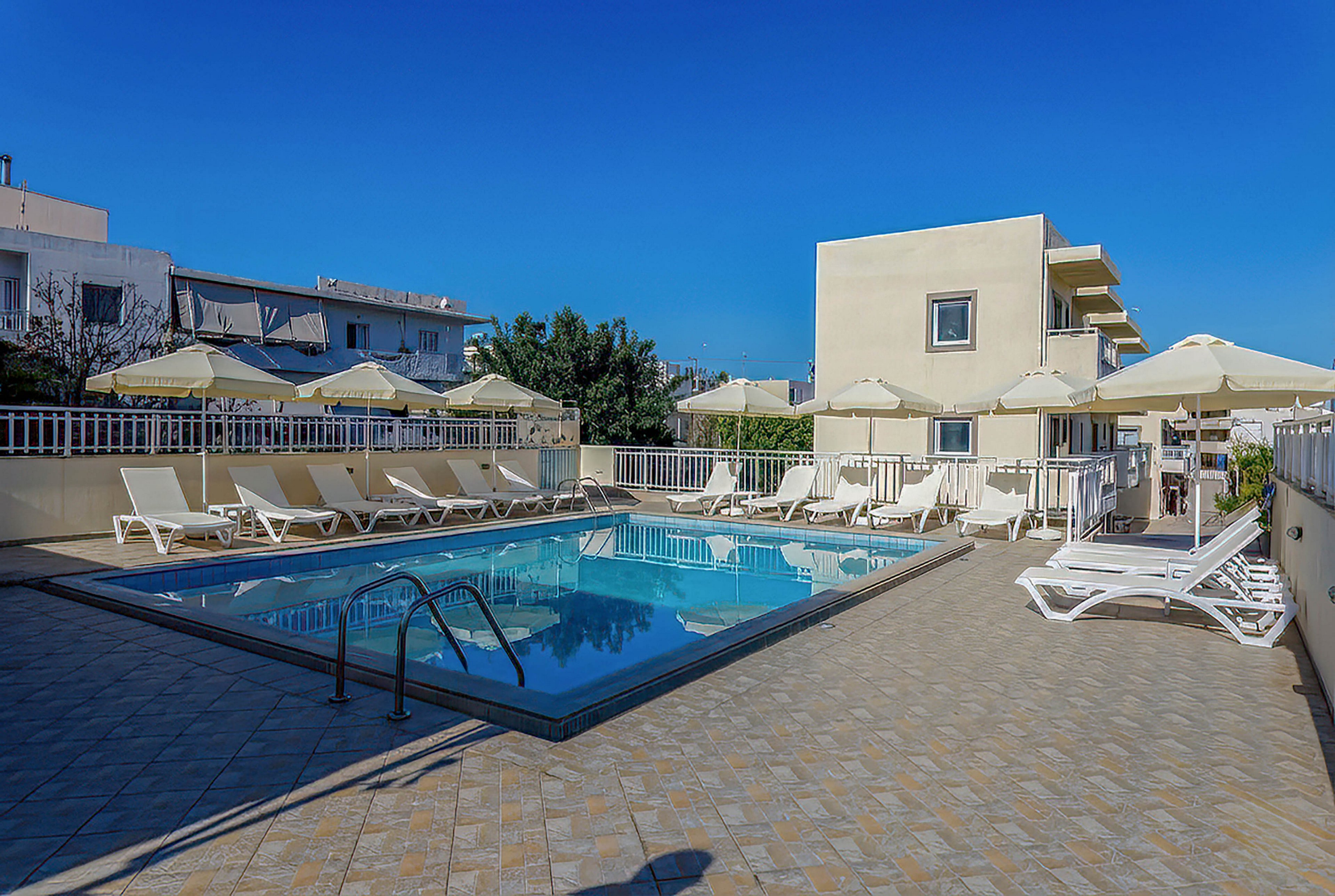 An outdoor swimming pool is surrounded by white lounge chairs and umbrellas in a modern hotel courtyard. The clear blue water reflects the bright sky, creating a calm and inviting atmosphere. A metal ladder leads into the pool, and a tiled deck provides ample space for sunbathing. Nearby low-rise buildings and greenery frame the scene, suggesting a warm vacation destination.
