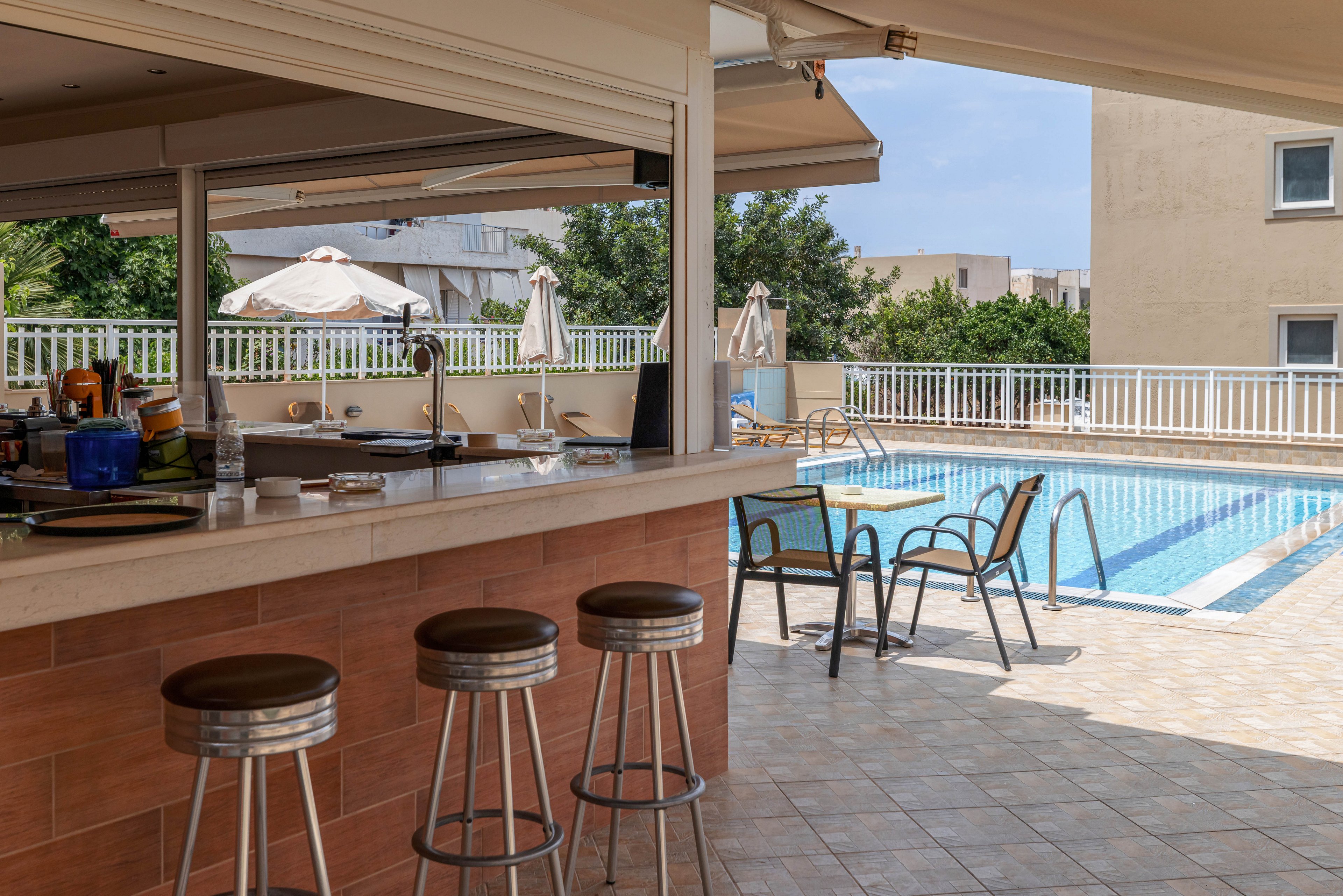 An outdoor bar area with metal stools overlooks a swimming pool in a sunny resort setting. The bar counter is clean and mostly empty, with a few dishes and utensils visible. Several lounge chairs and umbrellas surround the pool, suggesting a relaxed vacation atmosphere. Apartment-style buildings and greenery frame the background under a clear sky.