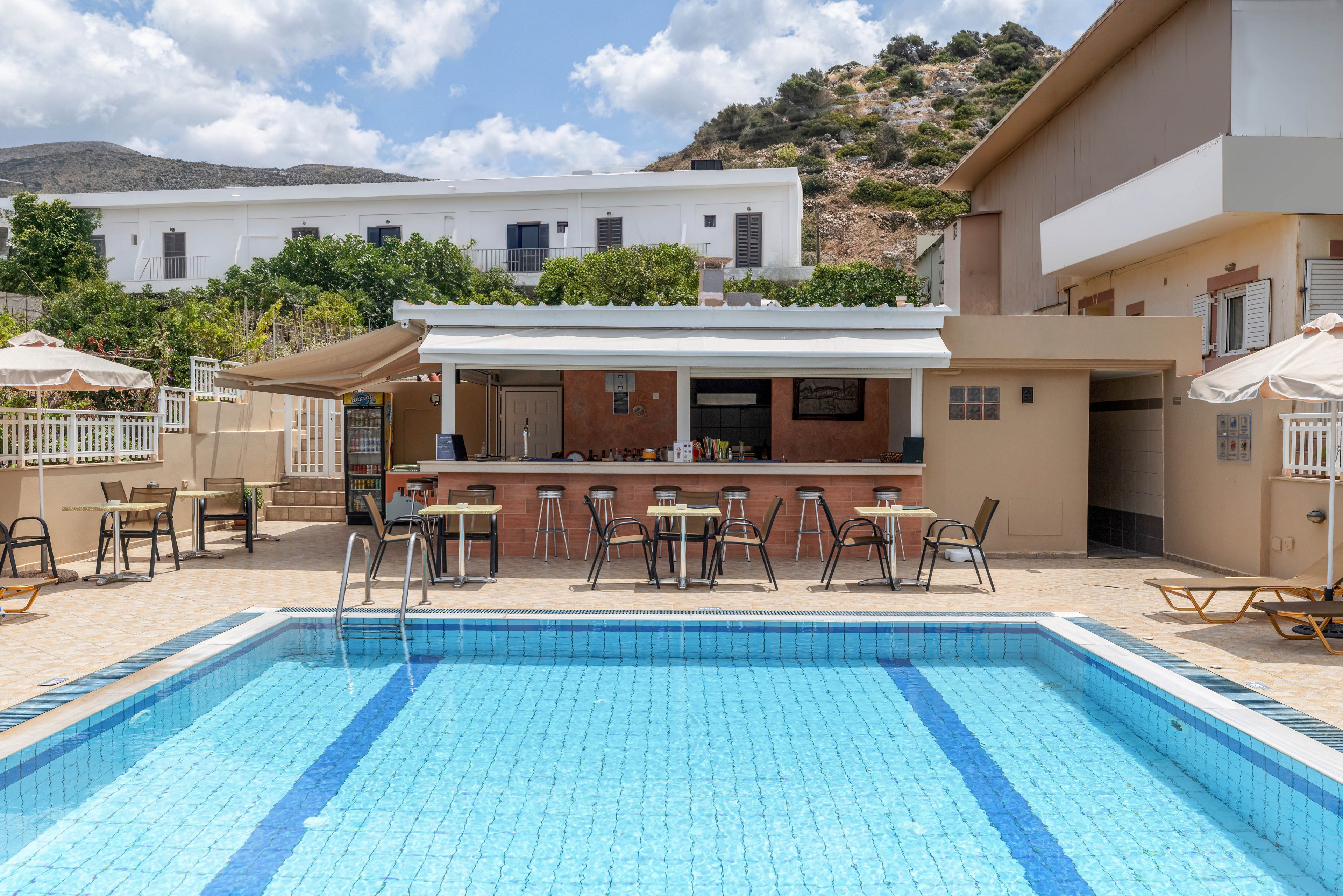An outdoor swimming pool is set in front of a small open-air bar with stools and tables. The scene appears to be part of a hotel or resort complex with low-rise buildings and balconies in the background. Sun loungers and umbrellas line the tiled pool deck, suggesting a relaxed vacation atmosphere. Hills and greenery rise behind the buildings under a partly cloudy sky.