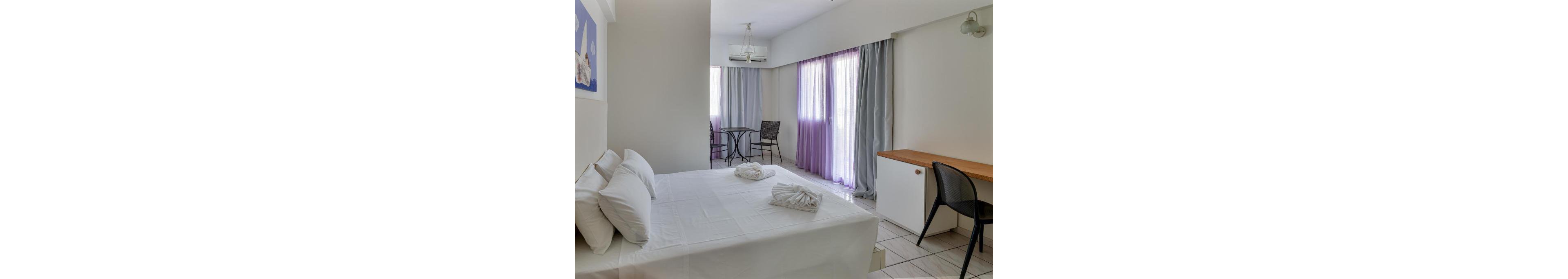A bright, minimalist hotel room features a neatly made double bed with white linens and folded towels. Floor-to-ceiling windows with sheer purple and gray curtains allow natural light to fill the space. A small round table with two chairs sits near the balcony area, while a simple desk with a wooden top and black chair lines the opposite wall. The overall atmosphere is clean, modern, and inviting, suitable for a relaxing stay.