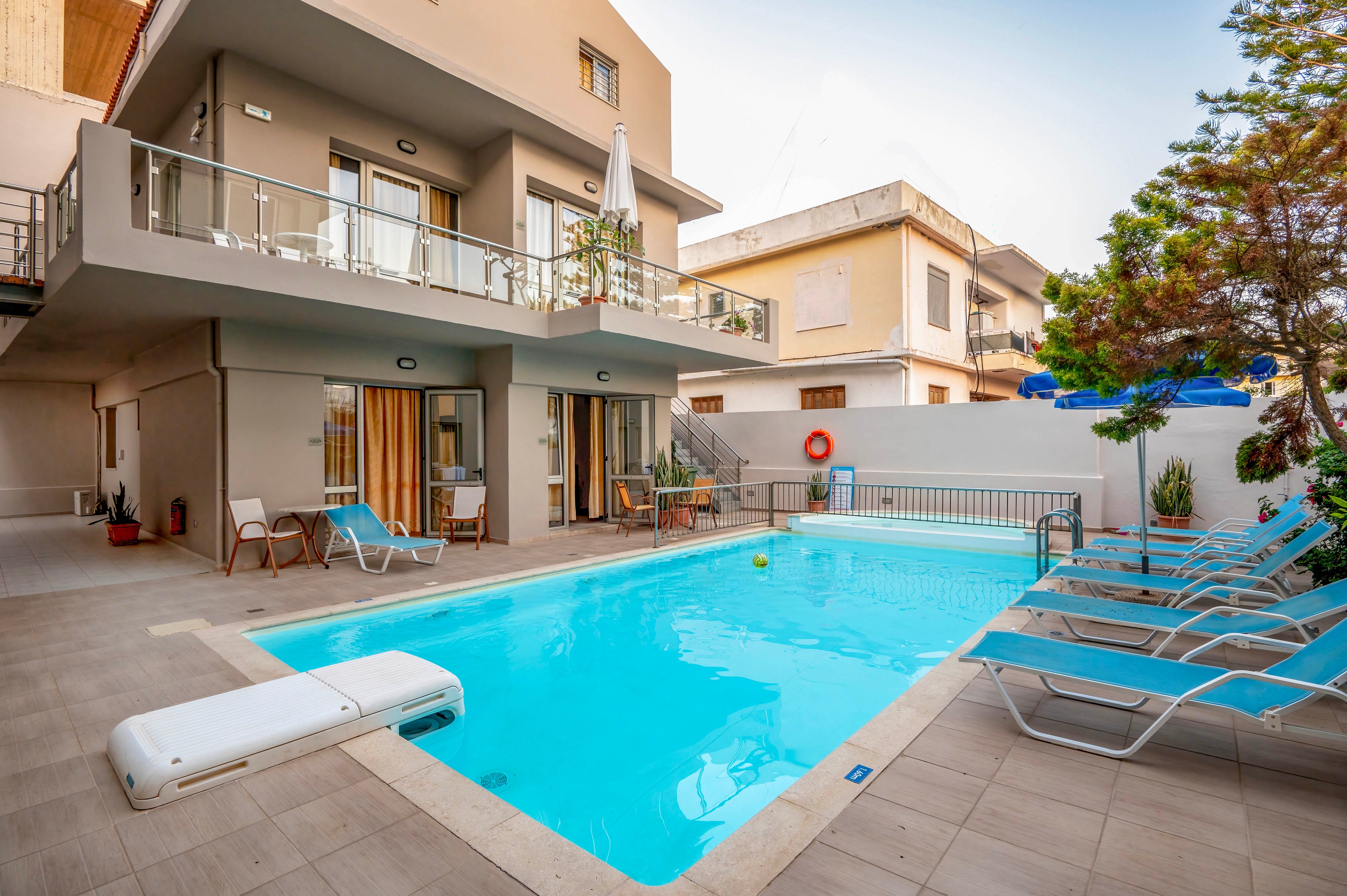 Contemporary two-story villa features a bright blue outdoor swimming pool in a residential neighborhood. Sun loungers with turquoise cushions line the tiled pool deck, creating a relaxing vacation atmosphere. Glass balconies, sliding doors, and neutral-colored walls give the building a modern, clean look. A lifebuoy, potted plants, and nearby umbrellas add subtle details to the tranquil setting.