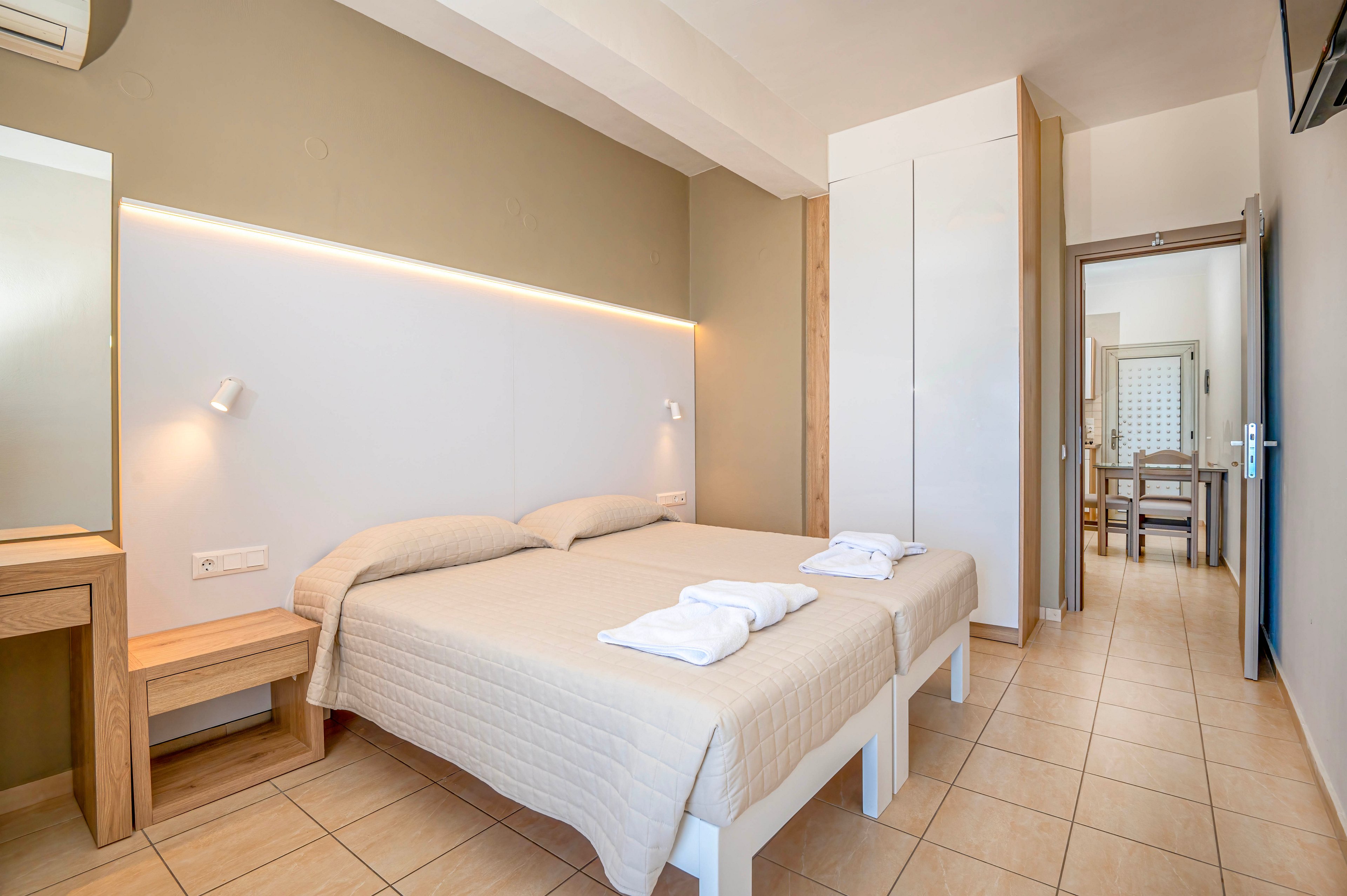 A clean, modern hotel room features two neatly made twin beds with folded white towels placed on top. The space has light neutral walls, tiled flooring, and simple wooden furniture including a desk and bedside table. Soft strip lighting above the headboard and small wall lamps create a bright yet cozy atmosphere. An open doorway leads to an adjacent sitting or dining area, enhancing the sense of space and comfort.