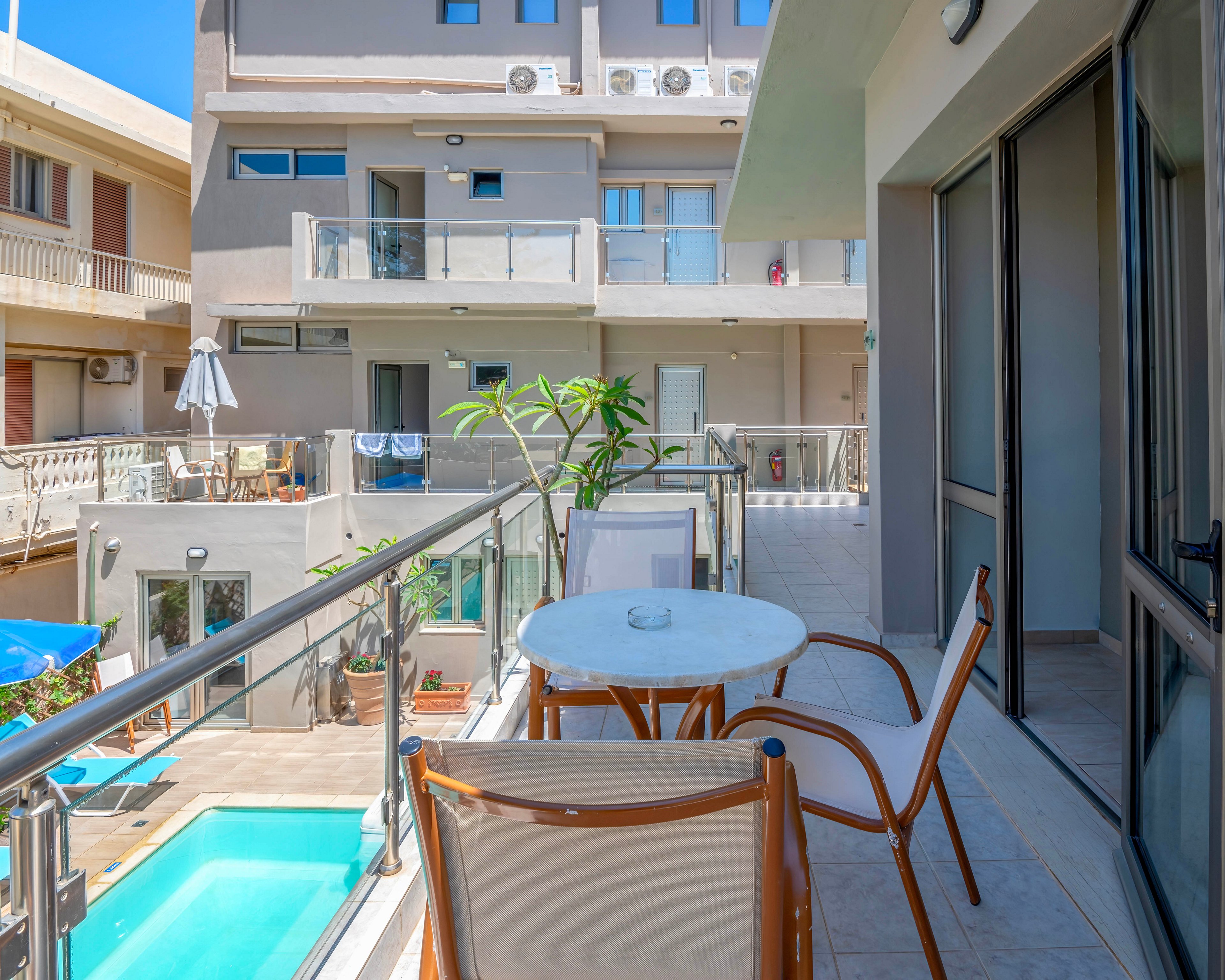 A bright hotel balcony features a small round table and two chairs overlooking a courtyard. Below, a rectangular swimming pool is surrounded by sun loungers and umbrellas. Modern apartment-style buildings with glass railings and multiple balconies frame the scene. The clear blue sky and potted plants add to the relaxed, vacation atmosphere.