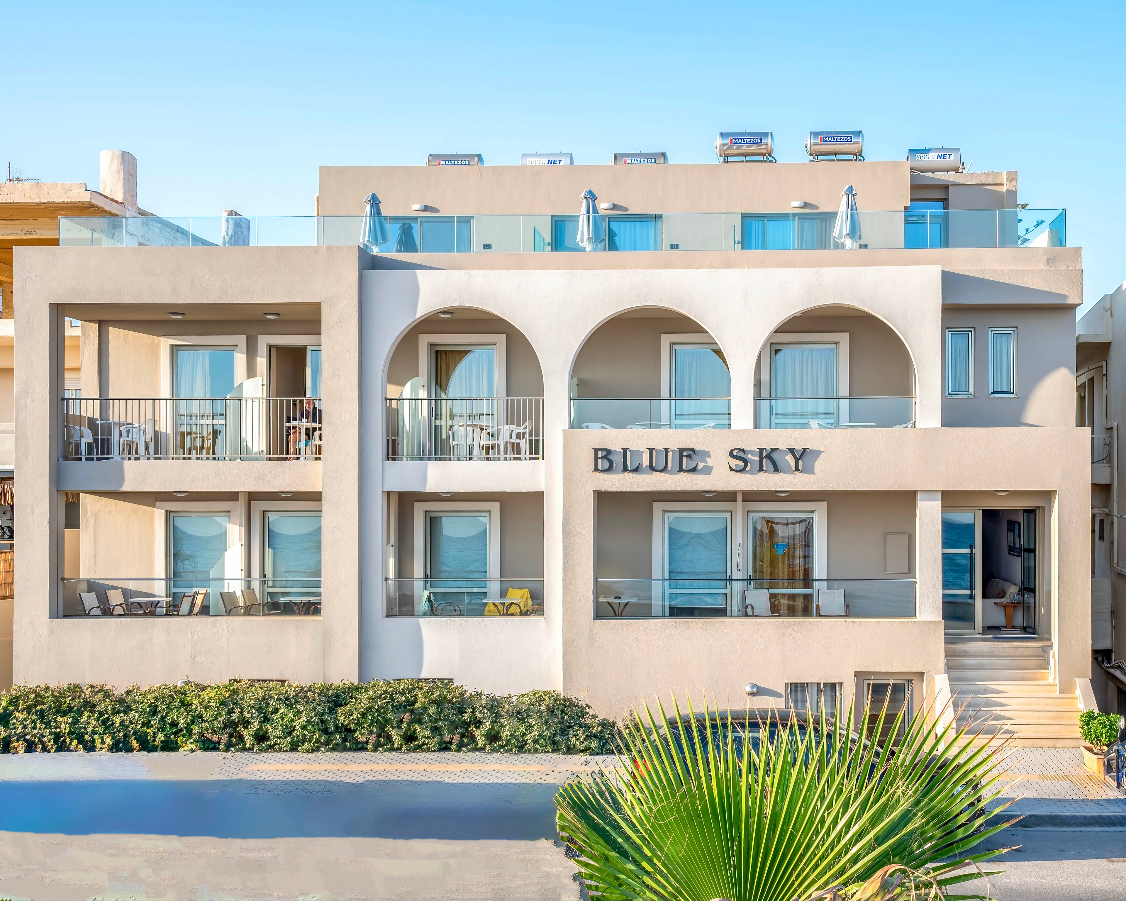 A contemporary multi-story hotel building features a symmetrical facade with large arches and private balconies. The name BLUE SKY is prominently displayed on the front of the structure. Sunlit terraces with outdoor seating overlook a neatly trimmed hedge and a palm plant in the foreground. The clear blue sky and bright daylight suggest a warm coastal or resort setting.