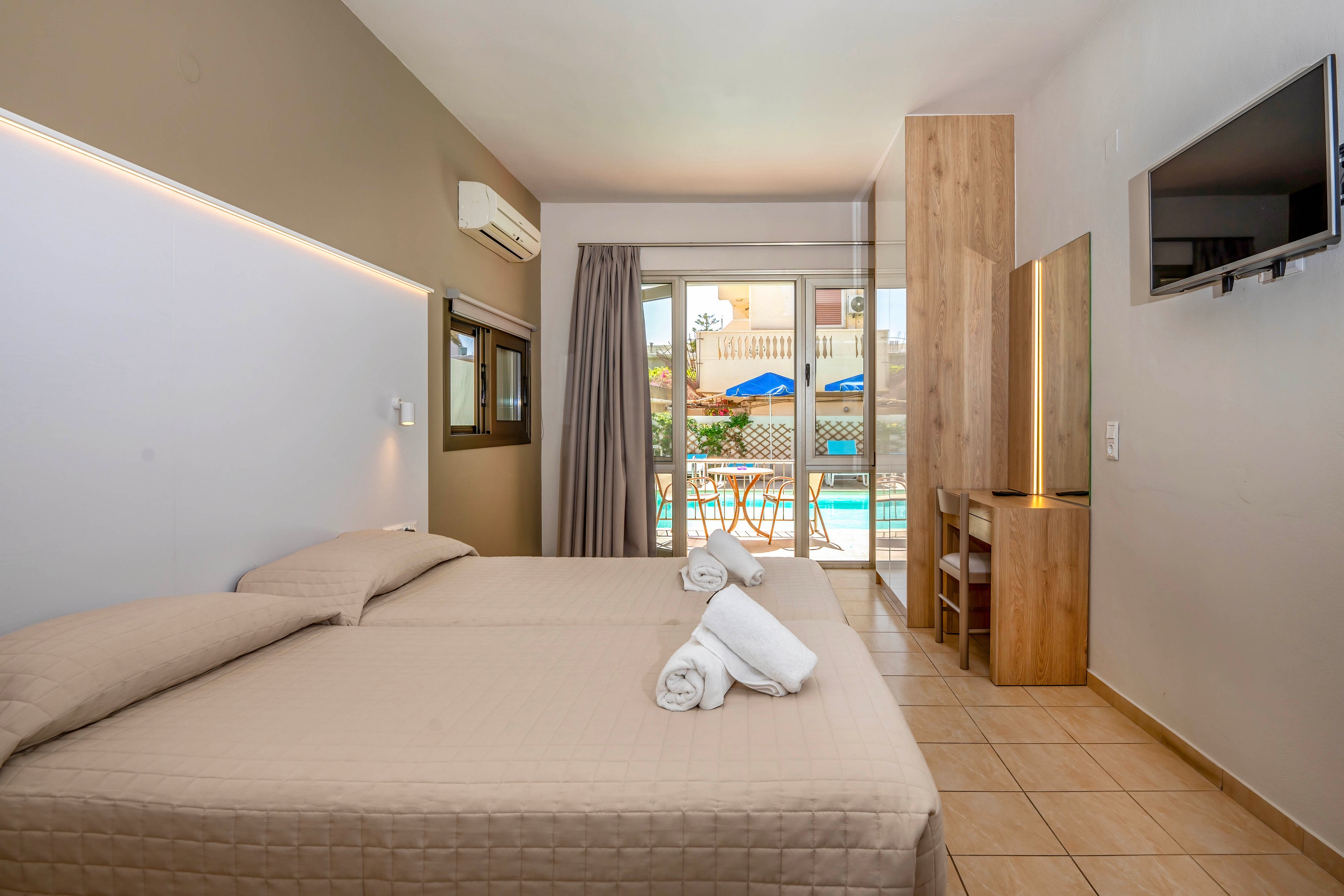 Modern hotel room featuring two neatly made twin beds with folded white towels on top. A wall-mounted television and simple wooden furniture create a clean, functional interior. Large glass doors open to a balcony overlooking a swimming pool area with blue sun umbrellas and loungers. Neutral tones and ample natural light give the space a calm, inviting atmosphere.