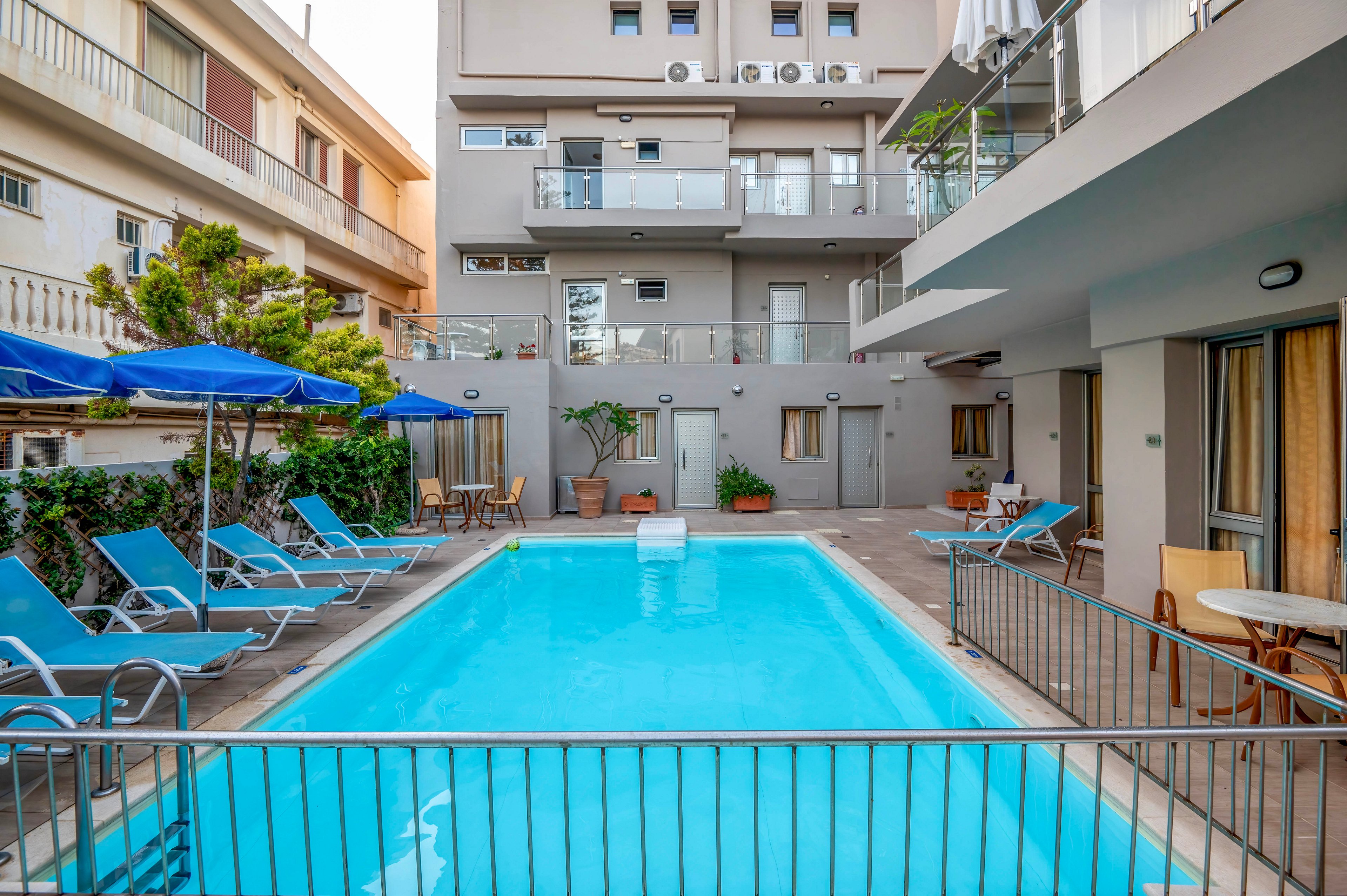 A rectangular outdoor swimming pool sits in the courtyard of a modern apartment or hotel building. Blue lounge chairs and umbrellas line both sides of the pool, creating a relaxed leisure area. The surrounding multi-story buildings feature balconies, windows, and potted plants. Metal railings frame the pool, and the overall scene suggests a quiet, urban vacation setting.