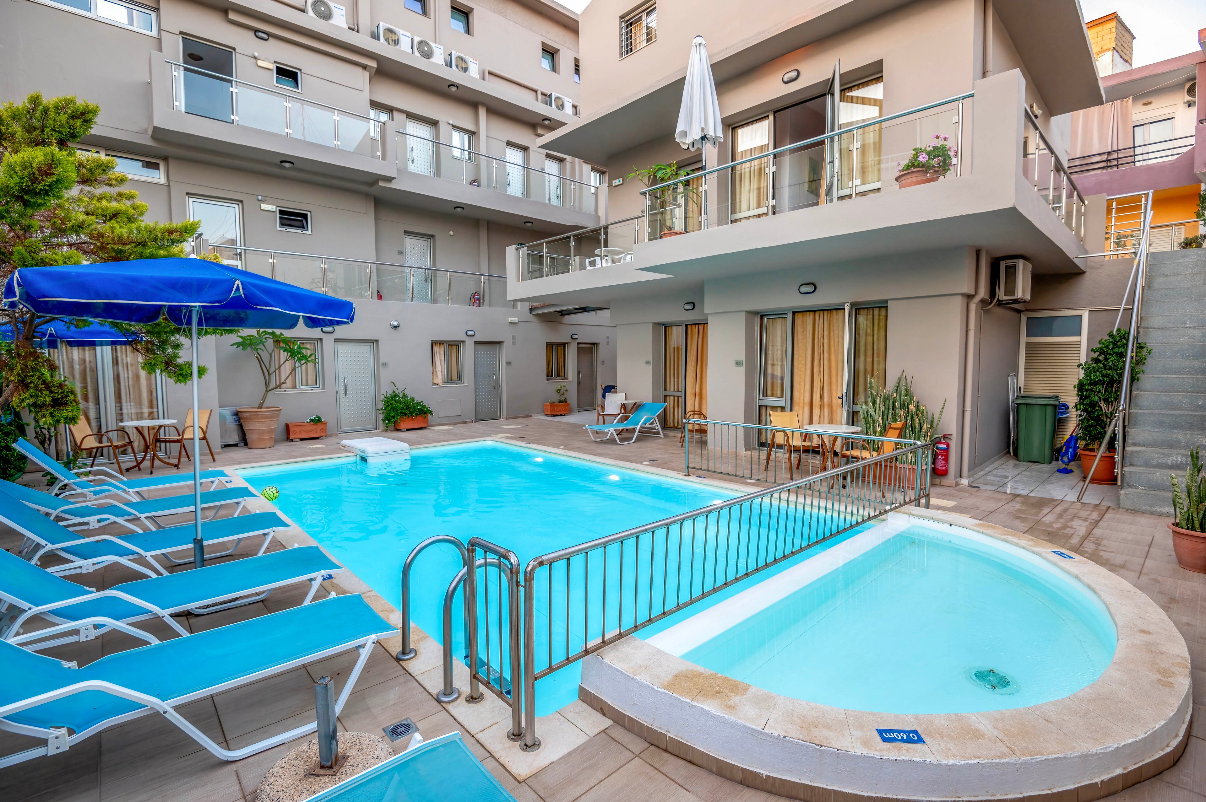 A small outdoor swimming pool and adjacent shallow pool sit in the courtyard of a modern apartment or hotel complex. Blue lounge chairs and umbrellas line the tiled deck around the water. Balconies with tables, chairs, and potted plants overlook the pool area, creating a cozy urban retreat. The scene is brightly lit, highlighting clean architectural lines and the inviting turquoise water.