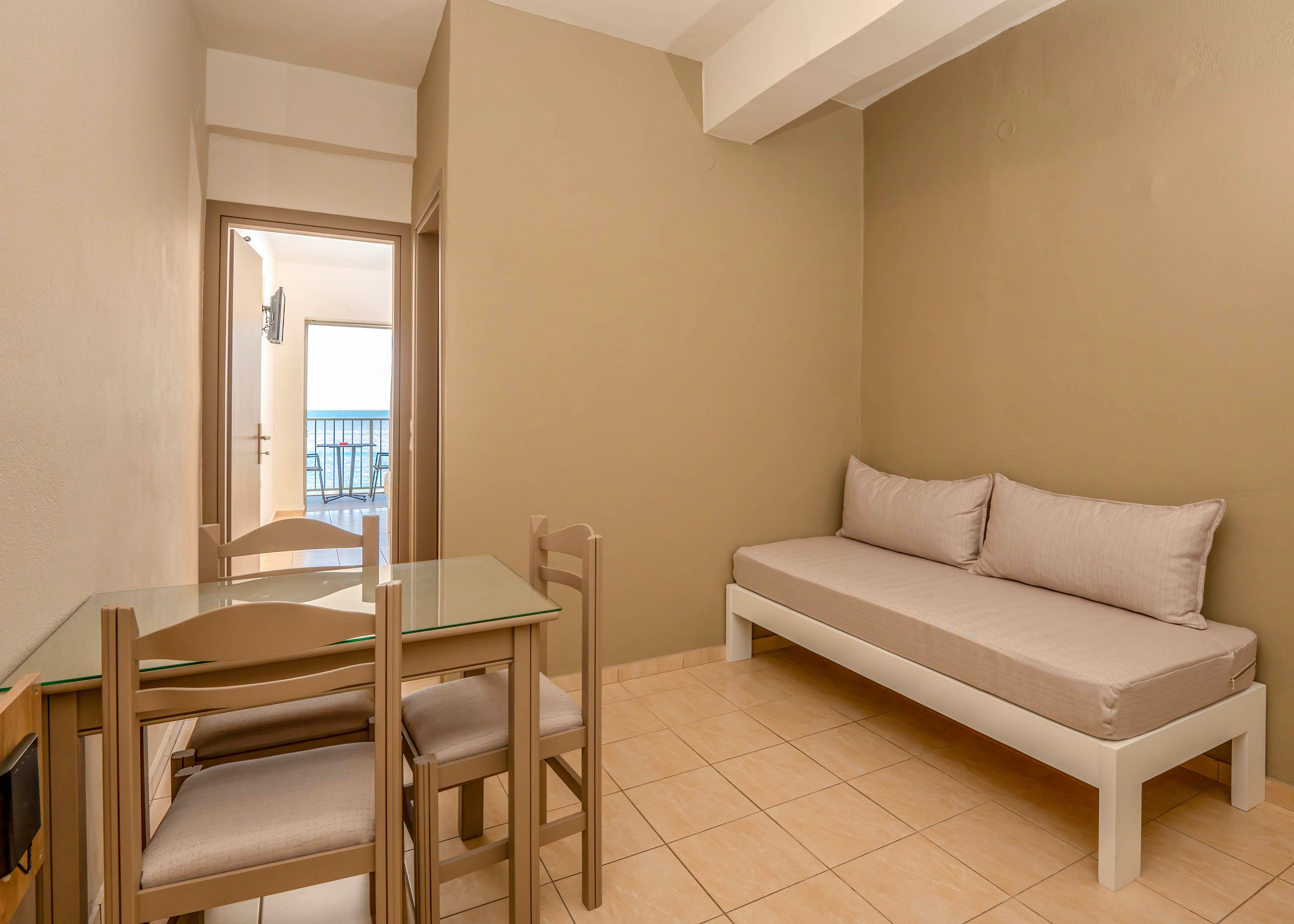 A compact hotel room features a simple beige daybed and a small glass-top dining table with wooden chairs. The space is finished with neutral walls and tiled flooring, creating a clean and minimalist look. Through the open doorway, a balcony with a railing and a view of the sea is visible, adding natural light and depth to the room. The overall atmosphere is calm and functional, suitable for short stays or vacation rentals.
