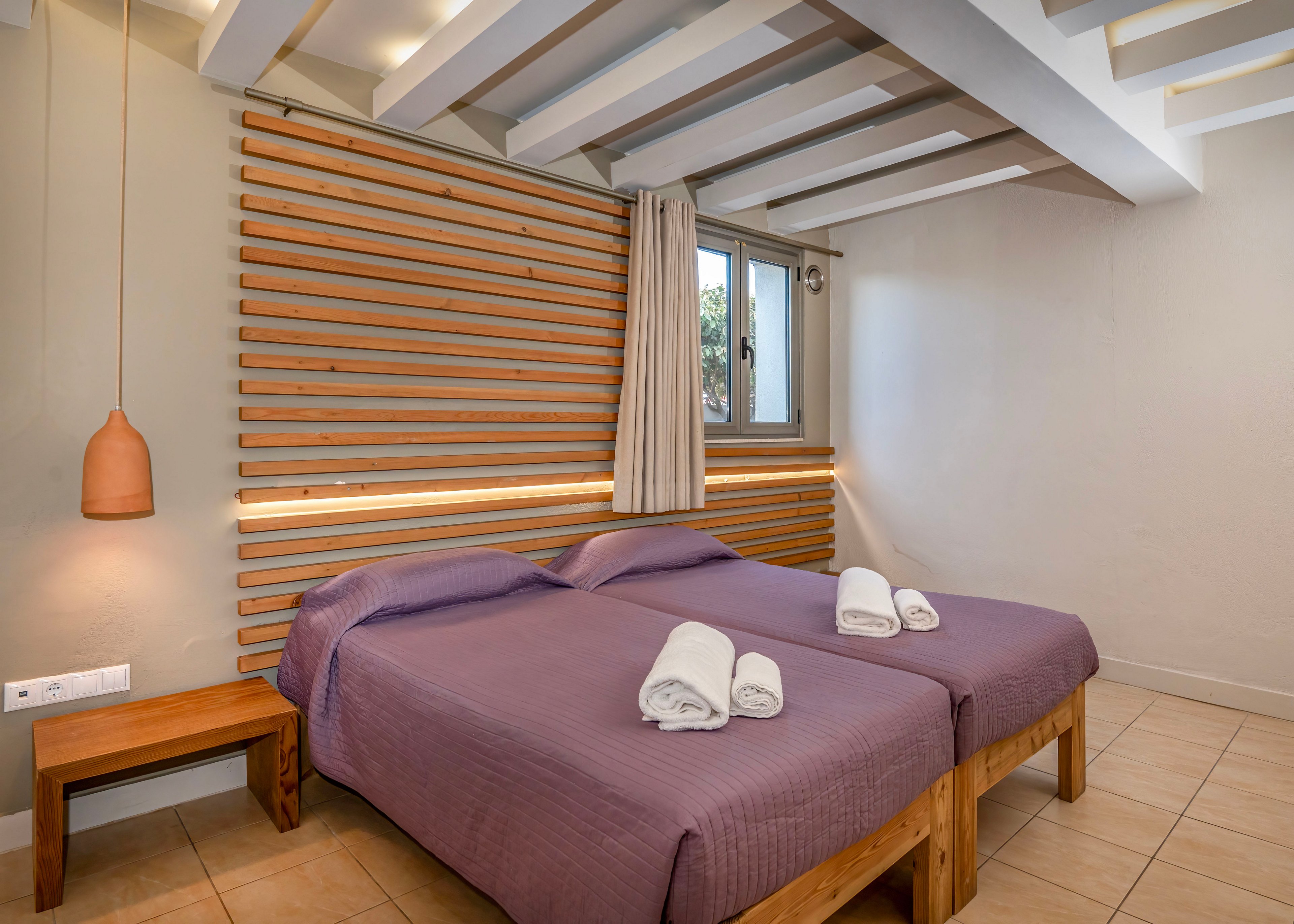 A contemporary bedroom features two twin beds with purple bedspreads and neatly folded white towels on top. The room has a wooden slatted headboard with integrated lighting and a small wooden side table. A single pendant light hangs beside the beds, and a window with beige curtains lets in natural light. The neutral walls and tiled floor create a clean, minimalist atmosphere.