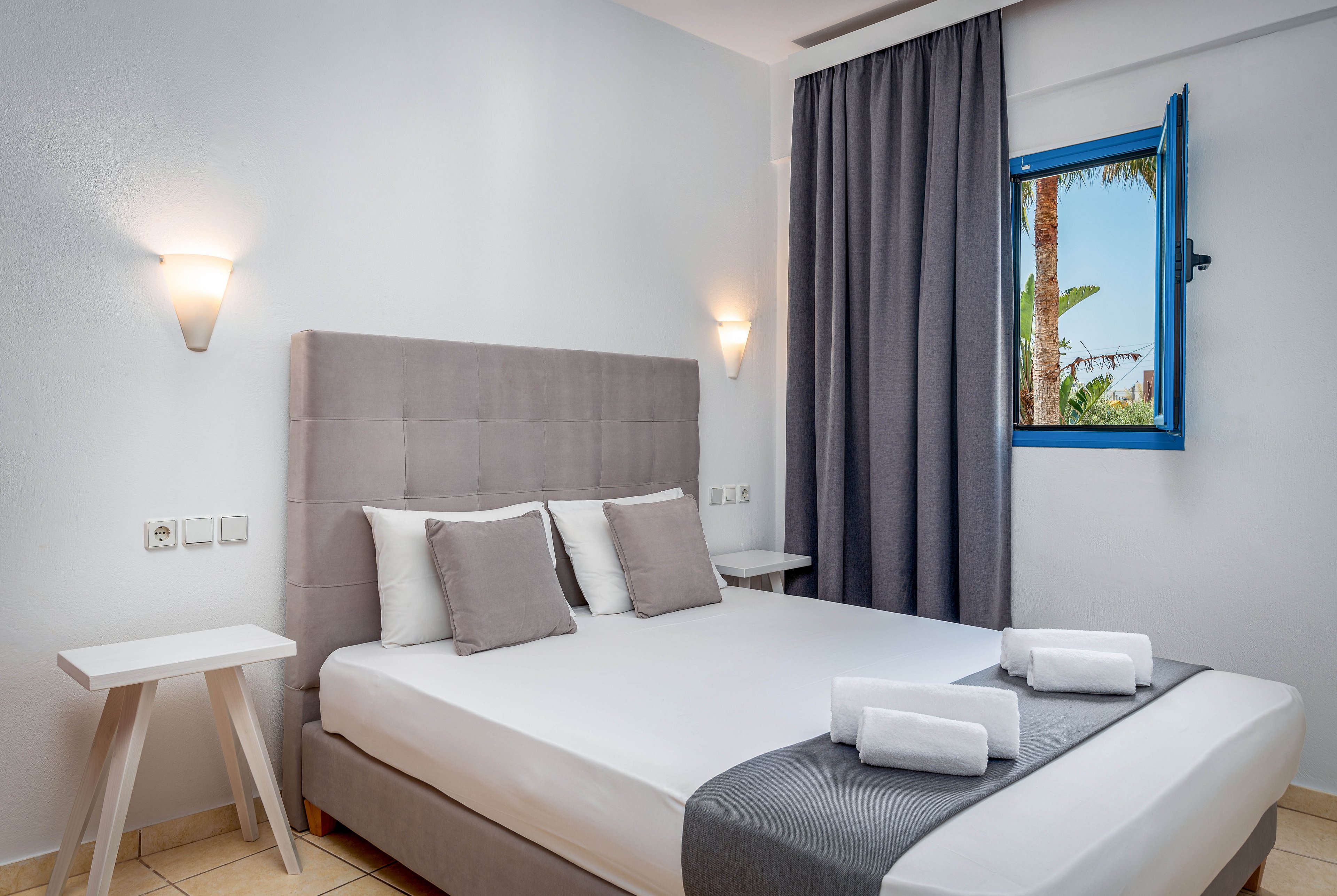 A neatly made double bed with a gray upholstered headboard is the focal point of this modern hotel room. White and gray bedding with rolled towels placed on top create a clean, minimalist look. Two small white bedside tables and wall-mounted lights add functional simplicity to the space. An open blue-framed window reveals palm trees outside, bringing natural light and a touch of greenery into the room.