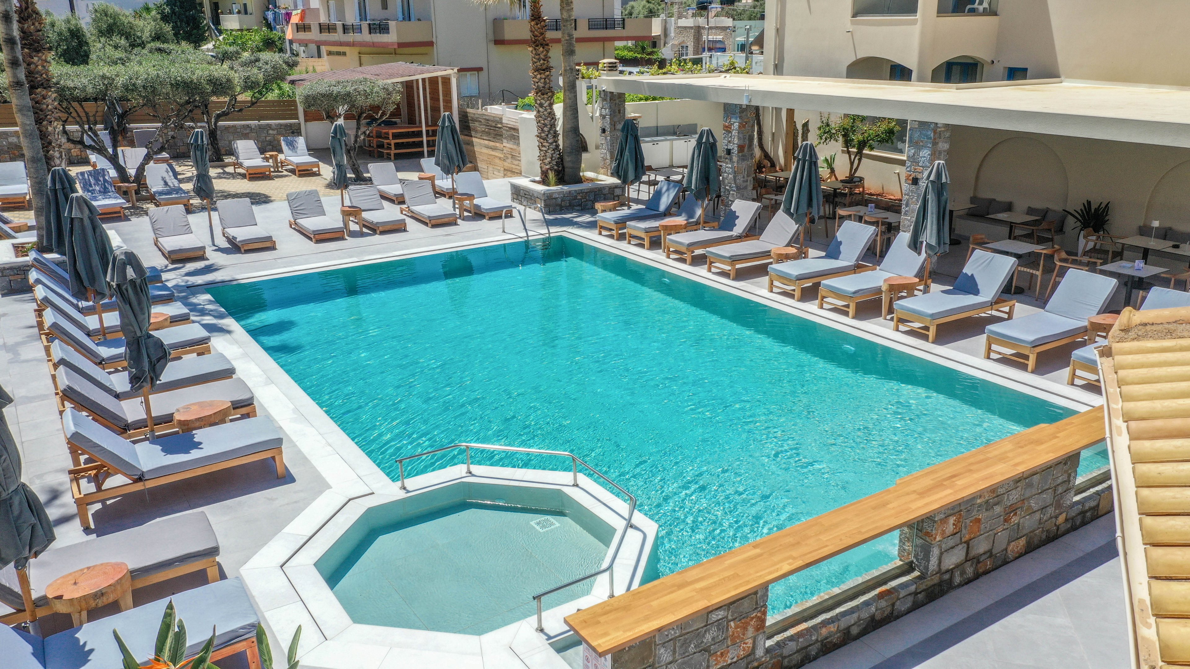 An outdoor resort swimming pool is surrounded by numerous sun loungers and umbrellas in a bright, sunny setting. A small adjacent hot tub or plunge pool sits in the foreground near stone walls and wooden railings. Modern buildings and palm trees frame the pool area, creating a relaxed vacation atmosphere. The clear blue water and neutral-toned furniture emphasize a clean, contemporary design.