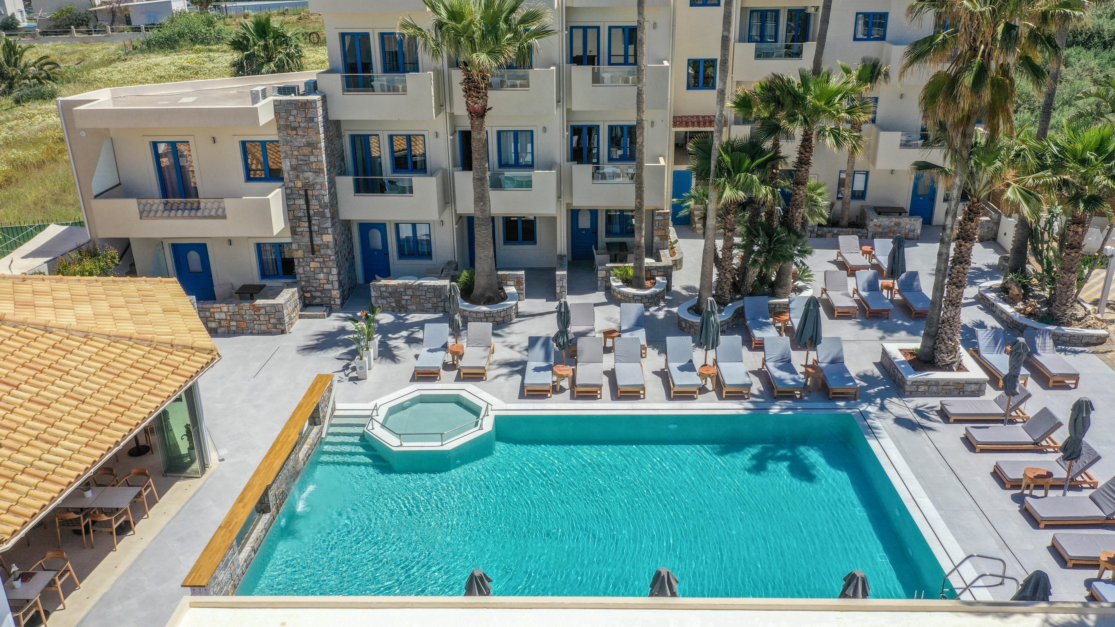 An aerial view shows a bright resort courtyard centered around a large turquoise swimming pool. Sun loungers and umbrellas line the poolside terrace, creating a relaxed vacation atmosphere. Palm trees and landscaped greenery add a tropical touch between the pool and the multi-story hotel building. A covered outdoor dining or bar area sits adjacent to the pool, suggesting leisure and hospitality. The scene is sunlit and inviting, emphasizing relaxation and warm weather.