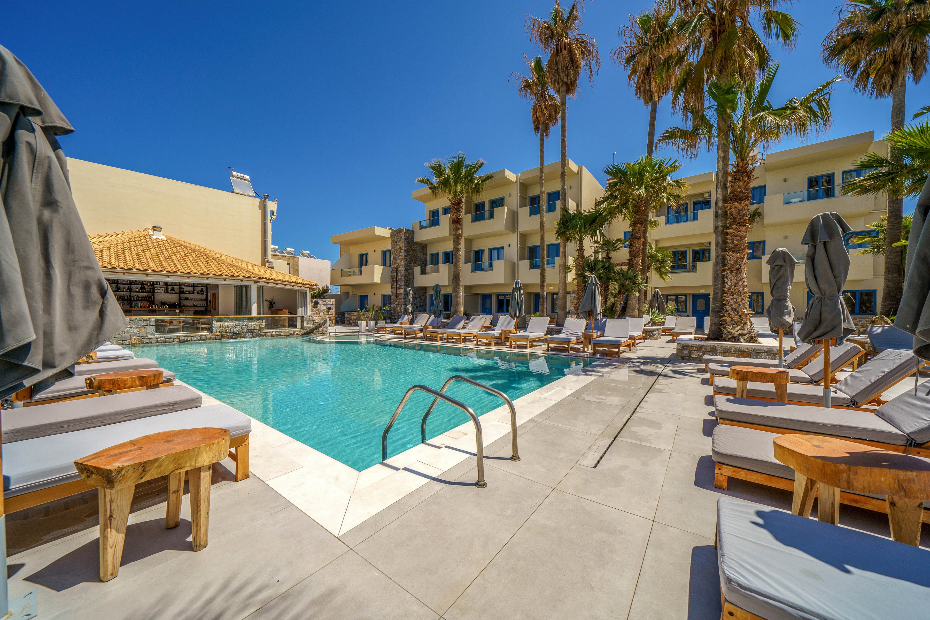 A bright resort swimming pool is surrounded by cushioned sun loungers and wooden side tables under a clear blue sky. Palm trees line the deck, adding a tropical feel to the modern multi-story hotel buildings in the background. Closed umbrellas and stainless steel pool ladders frame the water, suggesting a calm, relaxing atmosphere. The scene evokes a warm vacation setting focused on leisure and outdoor comfort.