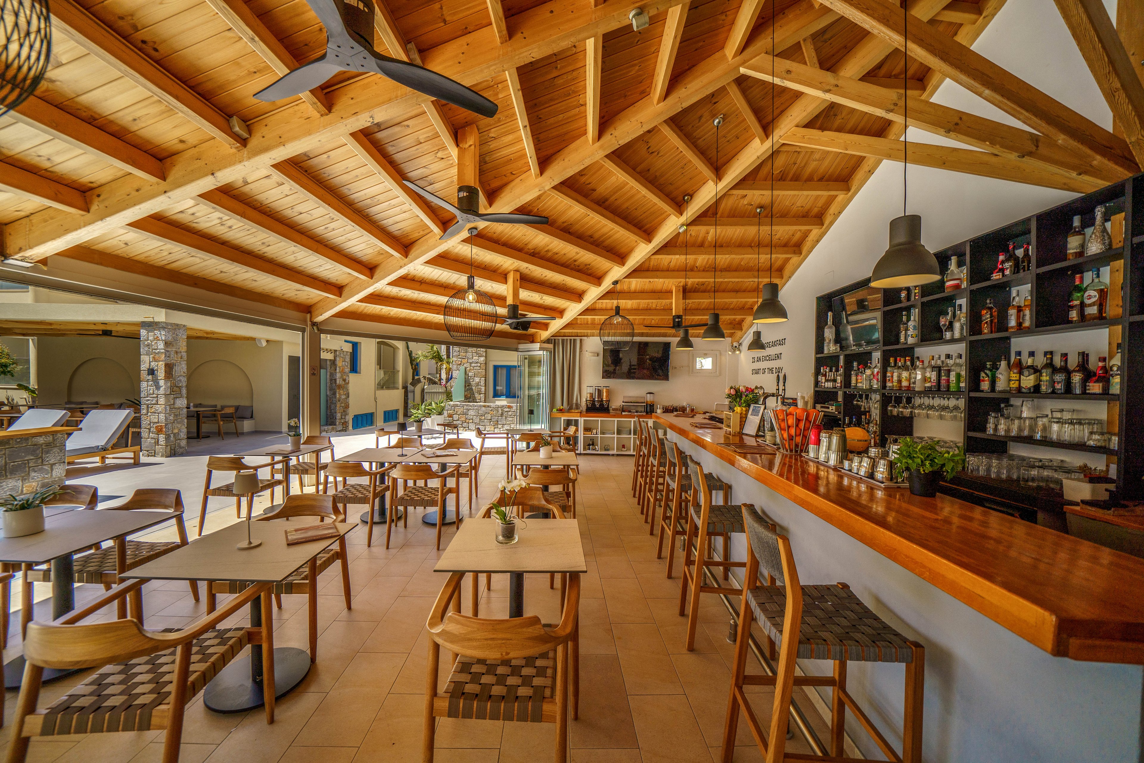 A contemporary bar and café interior features a long wooden counter and neatly arranged tables and chairs. The space has an open-air feel with large openings to the outside and abundant natural light. A wooden beam ceiling with ceiling fans adds warmth and architectural interest. Shelves behind the bar display various bottles and glassware, while small plants and décor create a relaxed, inviting atmosphere.