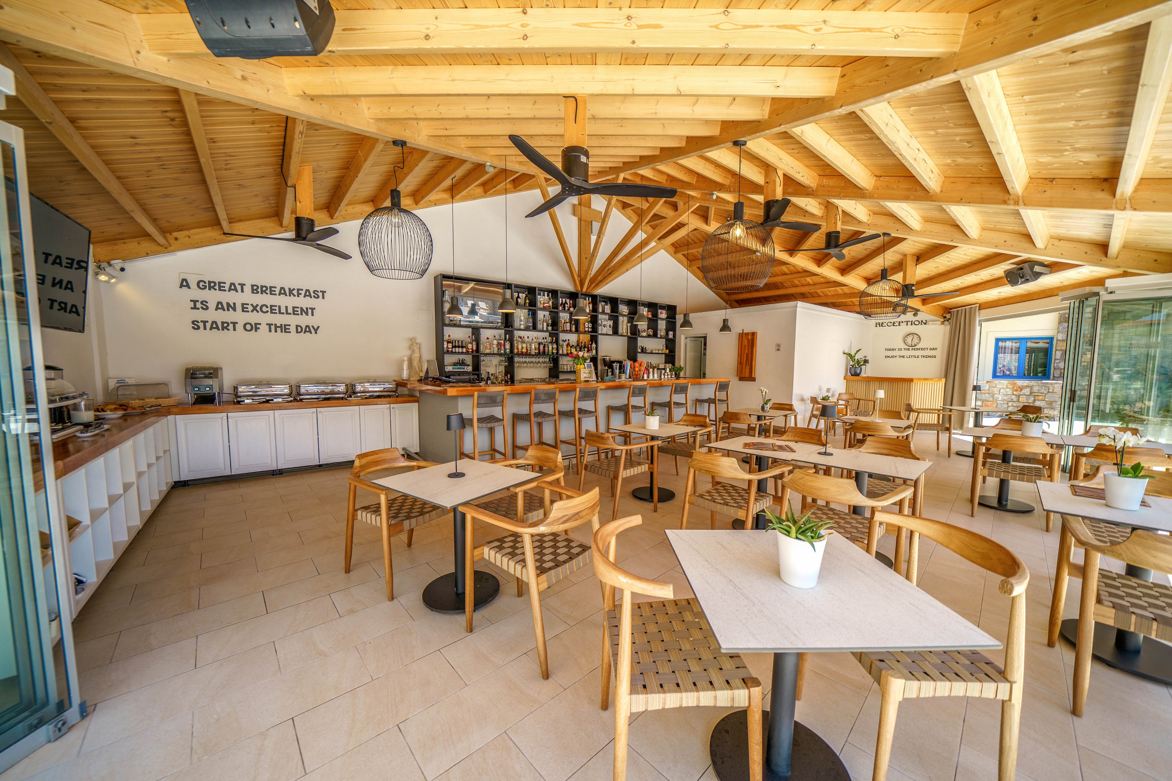 Spacious café interior featuring light wooden tables and chairs under a striking timber ceiling. The room is bright and airy, with large windows and an open layout leading to a counter and shelving filled with bottles and glassware. Wall text reads "A GREAT BREAKFAST IS AN EXCELLENT START OF THE DAY," emphasizing a welcoming atmosphere. Potted plants on the tables and neutral tones contribute to a clean, contemporary dining space.