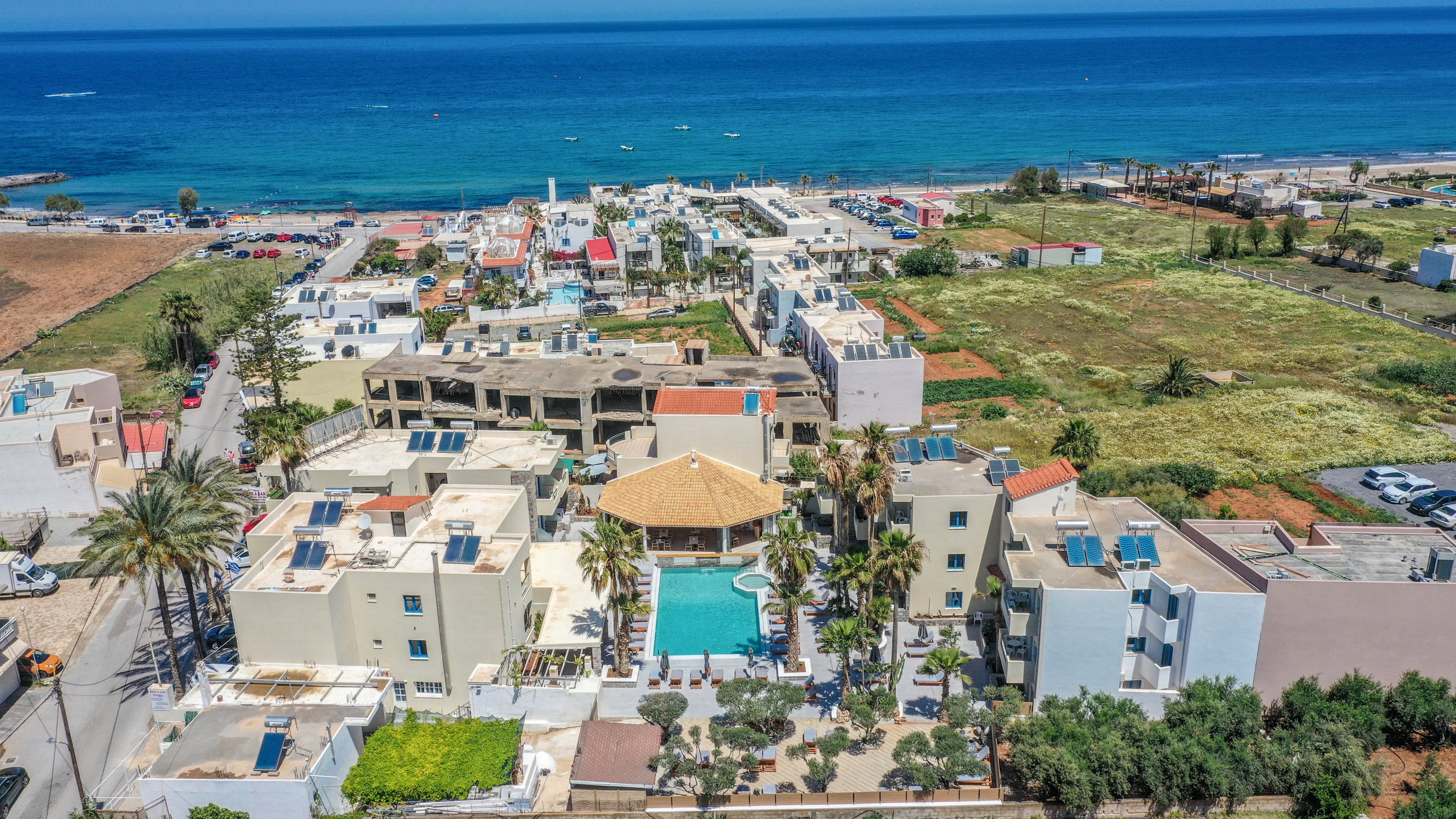 A large resort complex with multiple beige buildings and a central swimming pool is captured from above near a coastline. Palm trees and landscaped areas surround the pool, creating a tropical atmosphere within the property. Beyond the resort, open fields, roads, and additional low-rise structures extend toward the horizon. The blue sea and clear sky form a vivid backdrop, emphasizing the coastal vacation setting.
