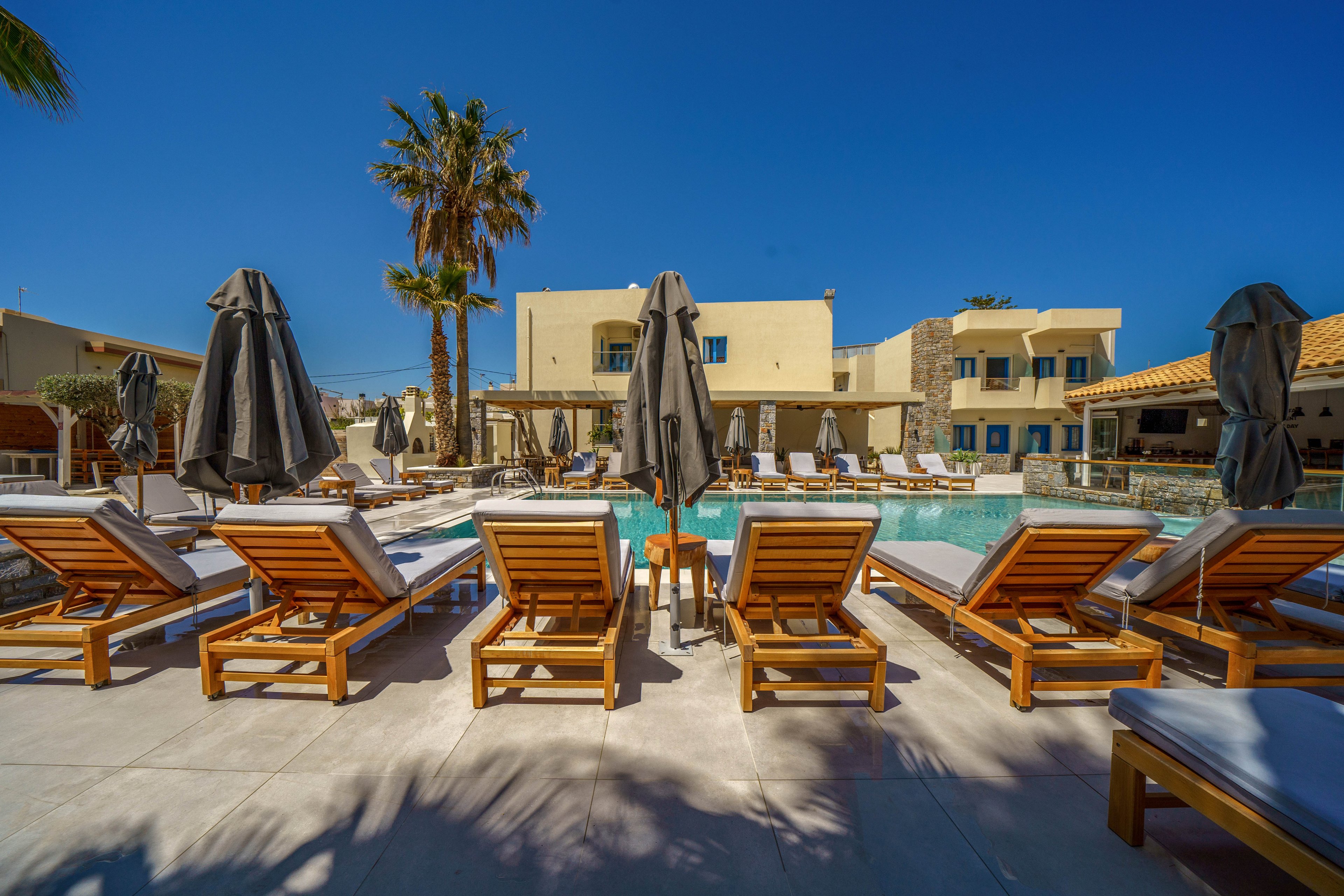 A row of wooden sun loungers with white cushions lines the edge of a clear blue swimming pool at a resort. Closed dark umbrellas stand between the chairs, suggesting a quiet, relaxing atmosphere. Beige low-rise buildings and balconies surround the pool area under a bright blue sky. A tall palm tree adds a tropical touch to the modern outdoor setting.