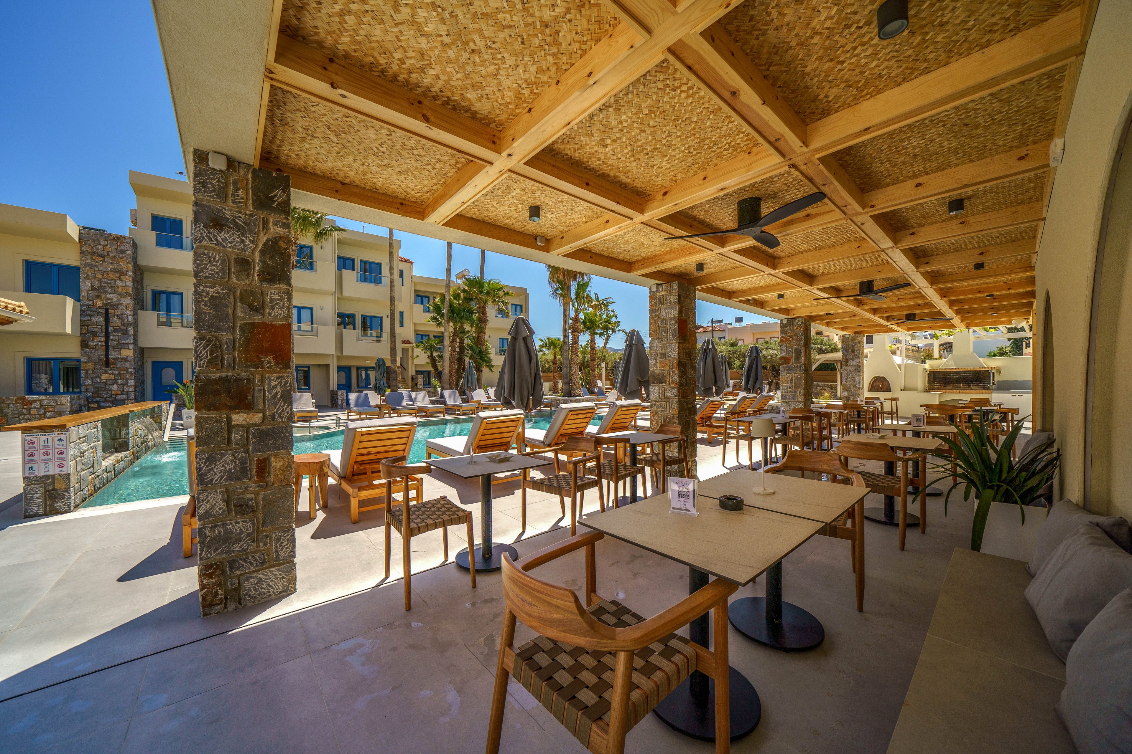 An open-air terrace with wooden pergola and tables faces a clear blue swimming pool. Sun loungers and closed umbrellas line the poolside, with modern resort buildings in the background. Stone columns and woven ceiling panels add a natural, Mediterranean-inspired touch. Bright daylight and palm trees create a relaxed vacation atmosphere.