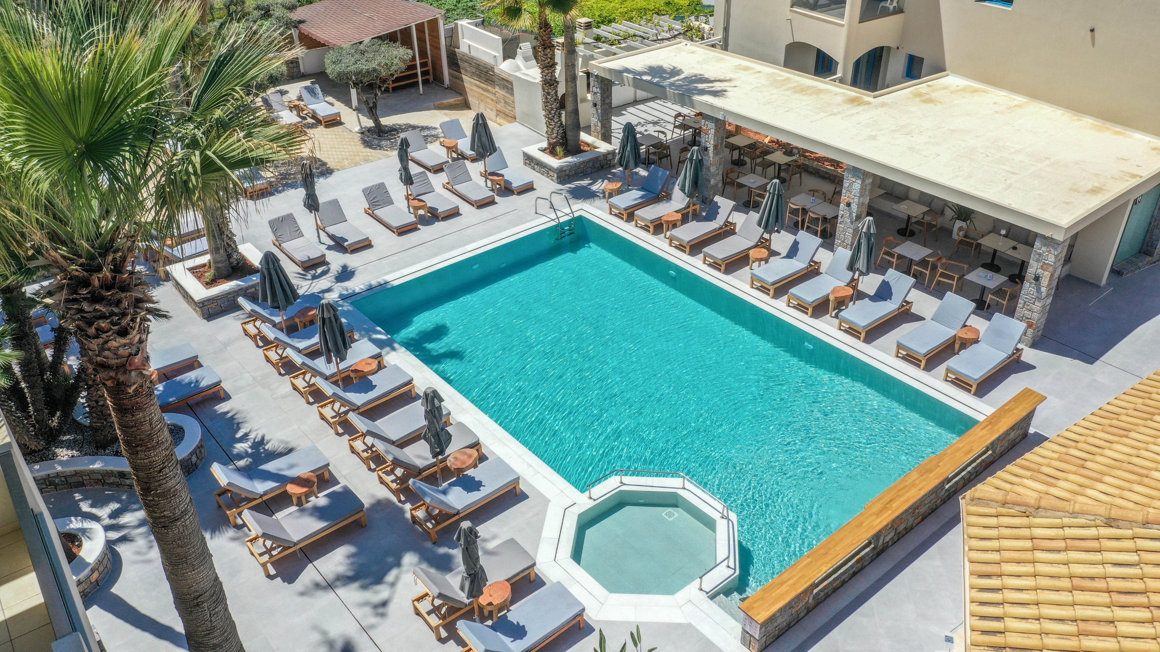 A large turquoise swimming pool is surrounded by numerous wooden lounge chairs and umbrellas in a resort courtyard. An adjacent small circular spa pool sits at one corner, bordered by a wooden railing. Palm trees and light-colored buildings frame the outdoor area, creating a relaxed vacation atmosphere. The scene is brightly lit, suggesting a warm, sunny day ideal for leisure and sunbathing.