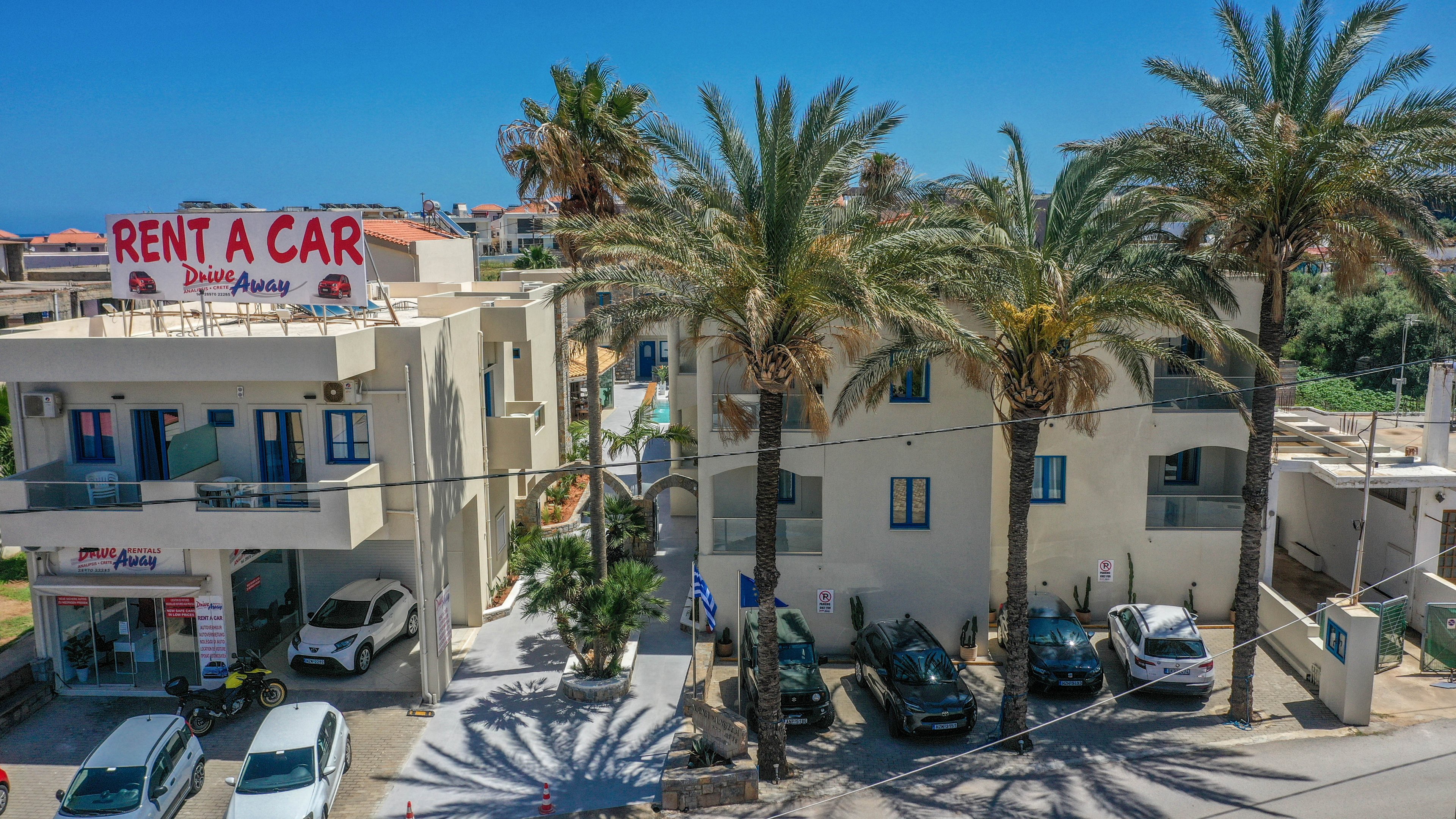 A rent a car office is situated in a light-colored apartment-style building along a sunny street. Several cars are parked in front of the property beneath tall palm trees, suggesting a vacation or tourist area. The sign on the rooftop clearly reads "RENT A CAR" with additional text below. Nearby buildings and a bright blue sky indicate a warm coastal town setting. Power lines and street details add to the everyday urban atmosphere.