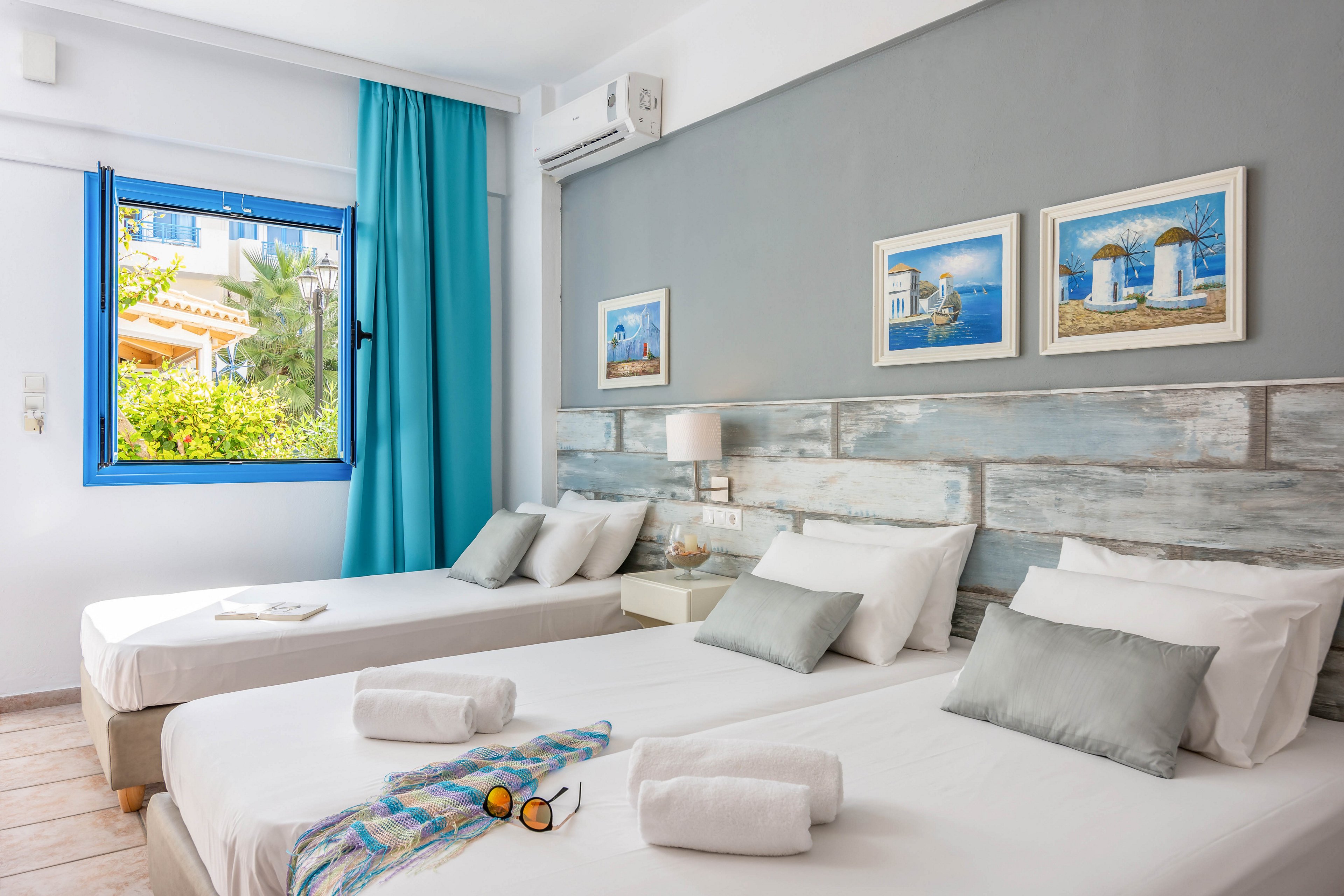 A light-filled bedroom features two neatly made single beds with white linens and soft gray accent pillows. The room has a coastal-inspired design, including distressed wood headboards and framed seaside artwork on a gray accent wall. A blue curtain and window frame add vibrant color, revealing greenery and buildings outside. Rolled white towels and a colorful striped scarf on the bed suggest a welcoming, vacation-ready space.