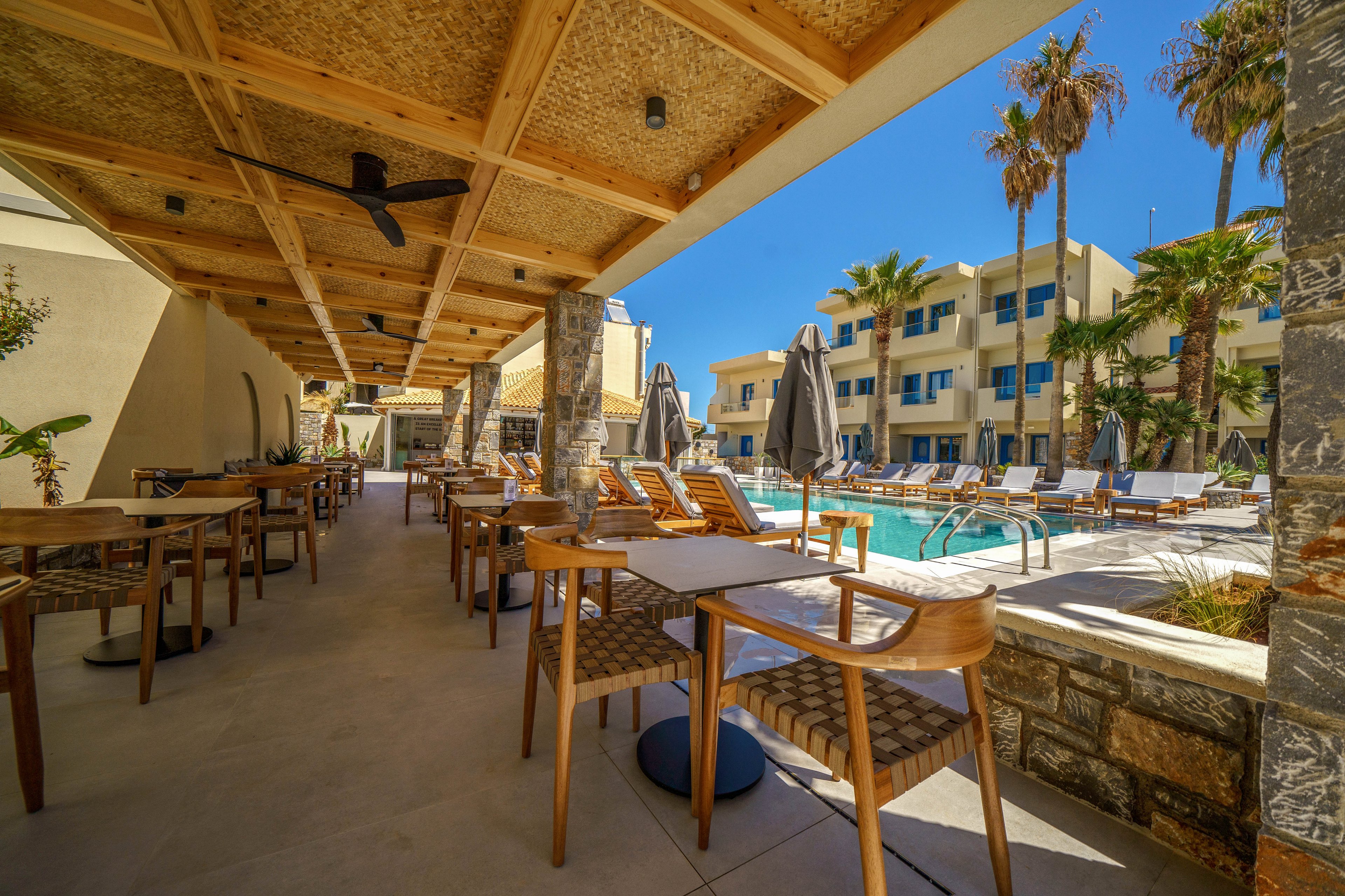 An open-air dining terrace with wooden tables and chairs faces a bright blue swimming pool. Sun loungers and large closed umbrellas line the poolside under a clear sky. Modern resort buildings and tall palm trees frame the scene, creating a relaxed vacation atmosphere. Warm wood tones contrast with the turquoise water and light stone surfaces.