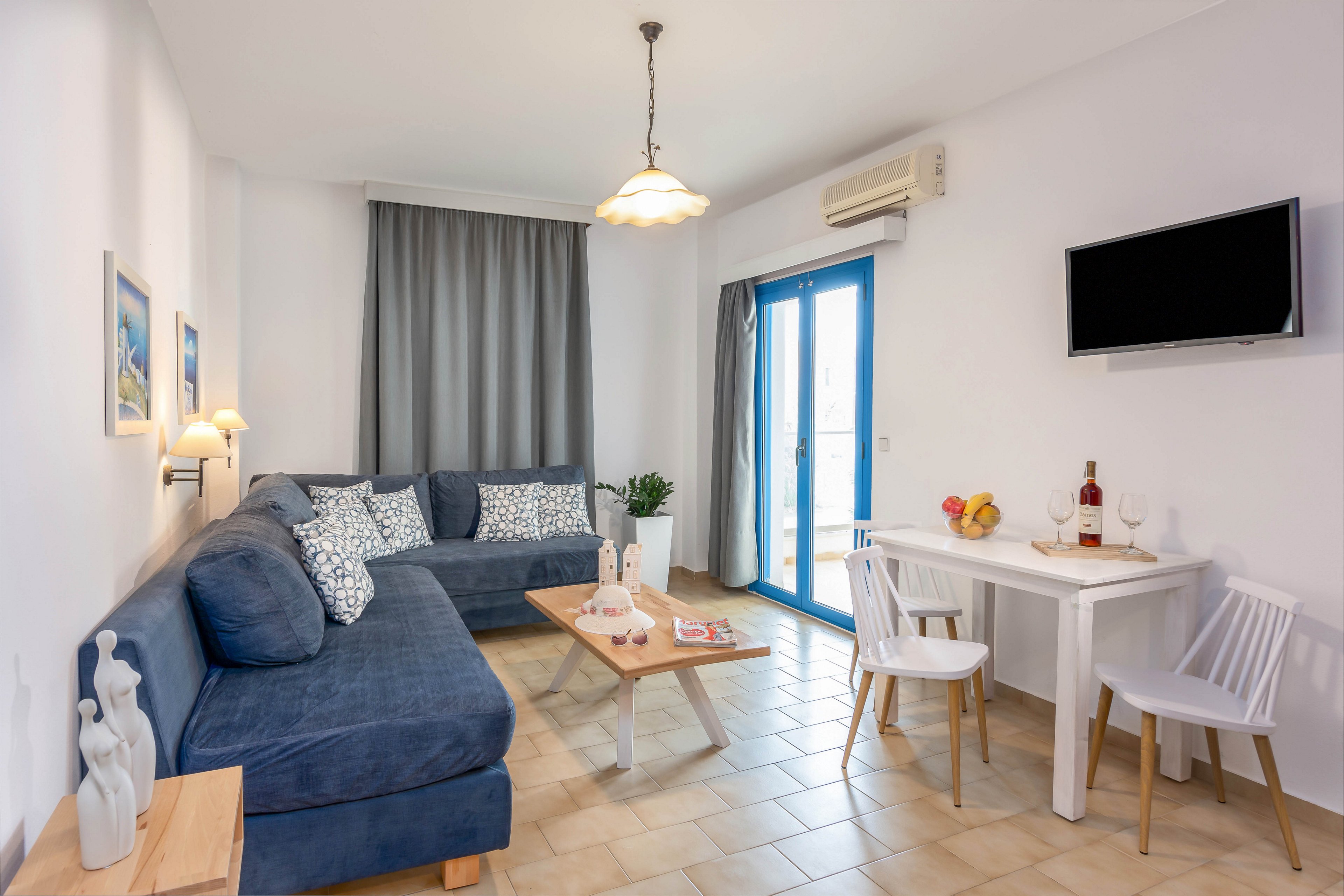 A modern living room features a large blue sectional sofa with patterned cushions and a light wood coffee table. The space opens to a small white dining table with four chairs, set with fruit and a bottle of wine. Blue-framed glass doors and a window with gray curtains bring in natural light, while a wall-mounted television and air conditioner complete the room. Neutral tiled flooring and white walls create a clean, airy, coastal feel.