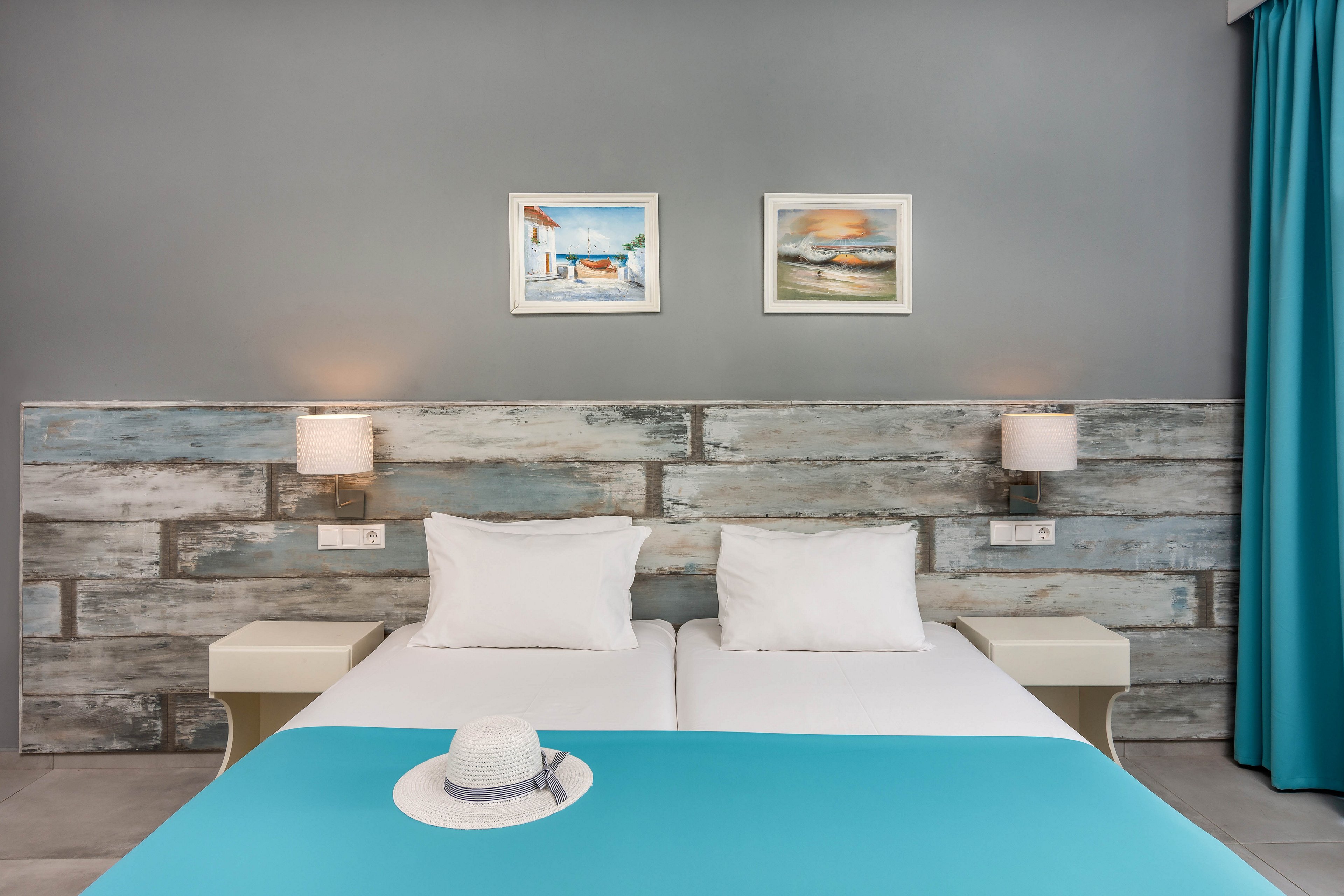 A neatly made double bed with white pillows and a turquoise bedspread stands against a rustic wood-paneled headboard. Two wall-mounted lamps flank the bed, with small bedside tables on each side. A white hat rests on the bed, adding a relaxed vacation feel to the scene. The gray wall features two framed coastal paintings, and a turquoise curtain completes the fresh, modern look.