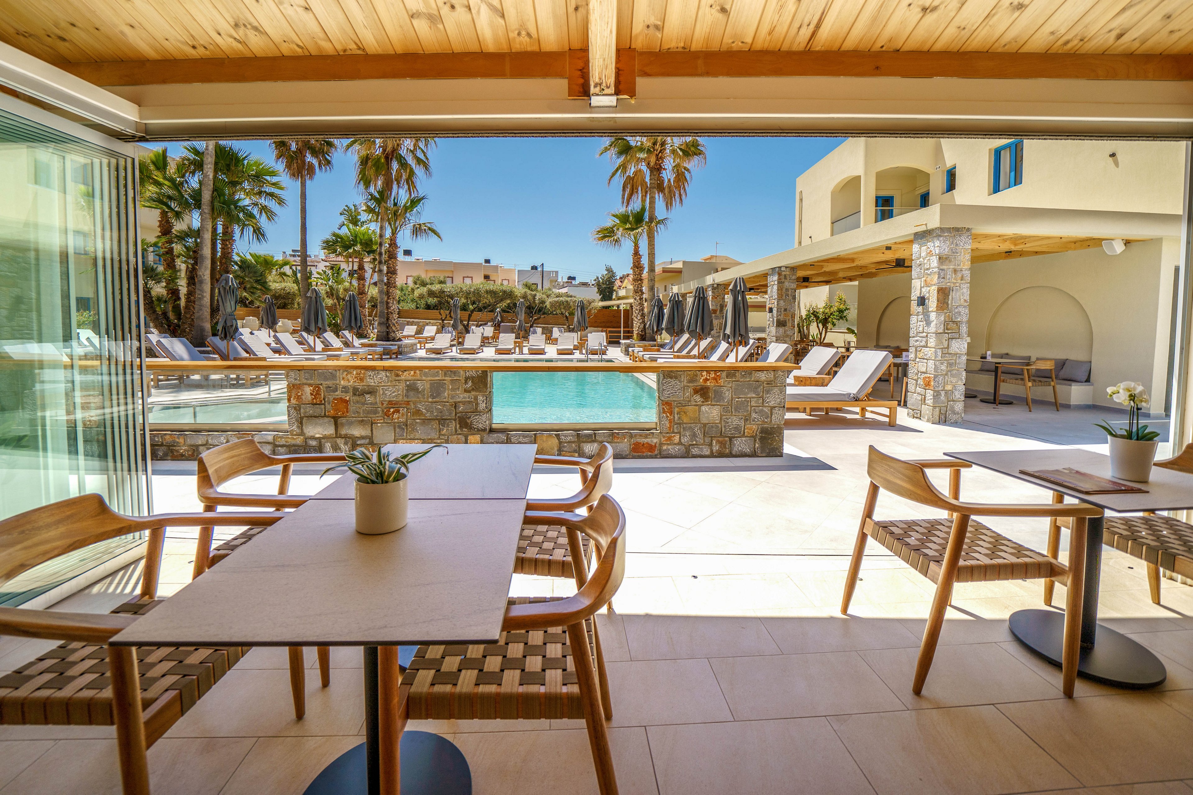 An open-air lounge area overlooks a bright resort swimming pool surrounded by sun loungers and umbrellas. Wooden tables and woven chairs sit under a shaded roof, creating a comfortable terrace for guests to relax or dine. Palm trees and stone walls frame the pool, while a modern hotel building rises in the background. The clear blue sky and warm light suggest a sunny vacation destination with a calm, inviting atmosphere.