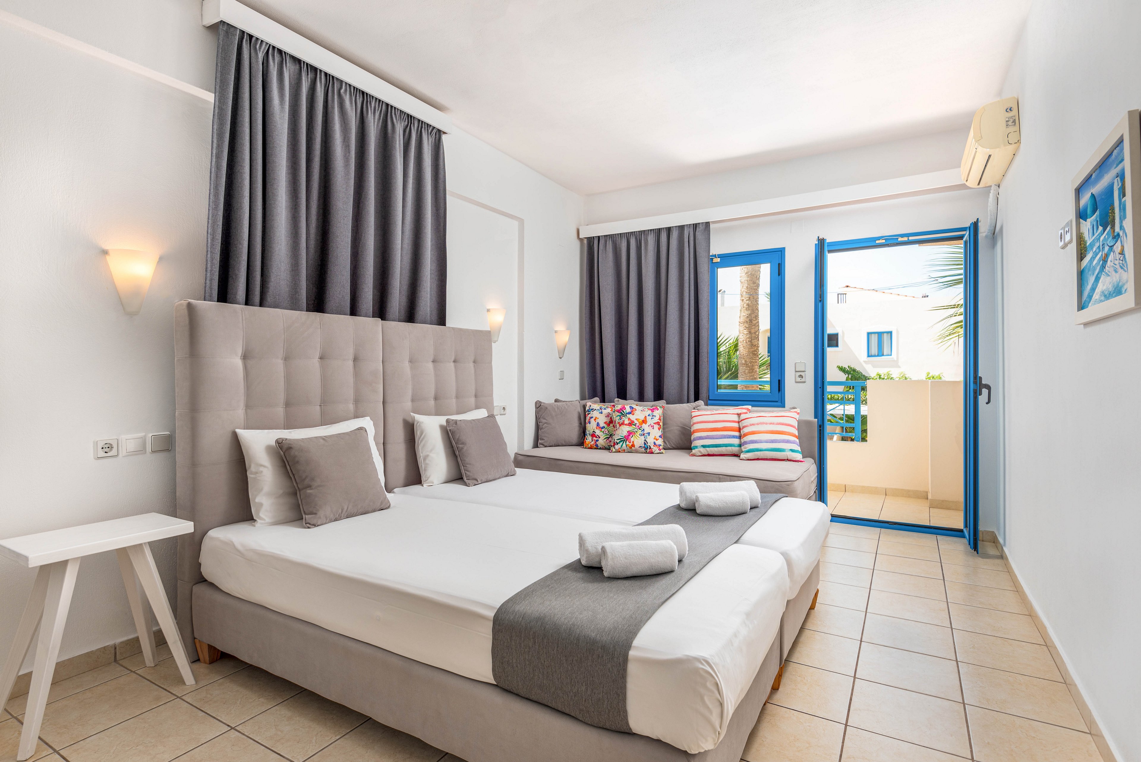 A modern hotel room features two neatly made twin beds with gray runners and folded white towels. A cushioned daybed with colorful patterned pillows sits against the wall beside the beds. Light beige floor tiles, white walls, and dark gray curtains create a clean, contemporary look. Blue-framed windows and balcony door open to an outdoor view, adding a fresh, coastal feel to the space.