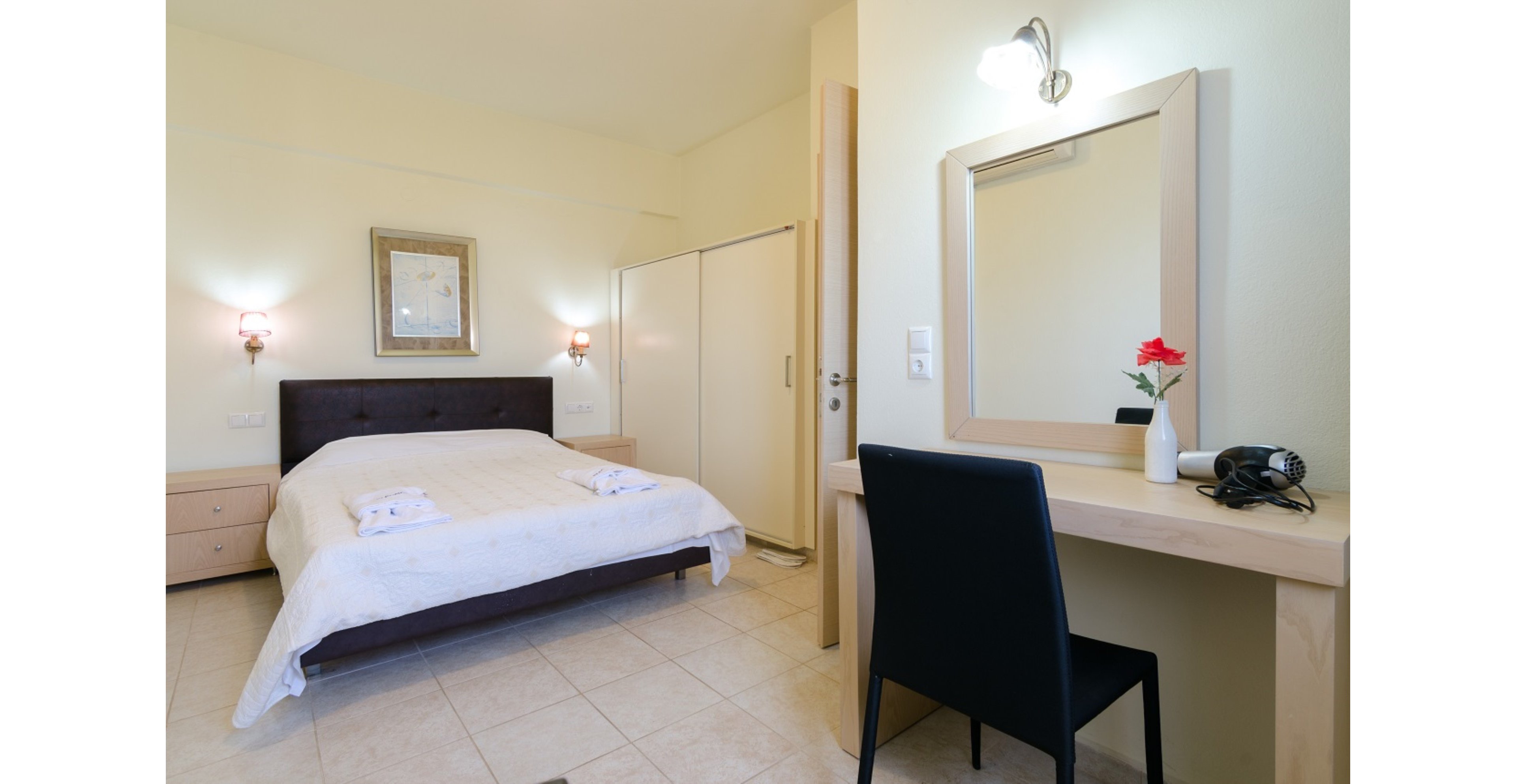 A tidy bedroom features a double bed with folded towels placed neatly on top. The room includes light wood bedside tables, a wardrobe, and a simple desk with a black chair. Above the desk is a large mirror, a wall light, and a small vase holding a single red flower. Soft, neutral colors and clean lines create a calm, modern atmosphere.