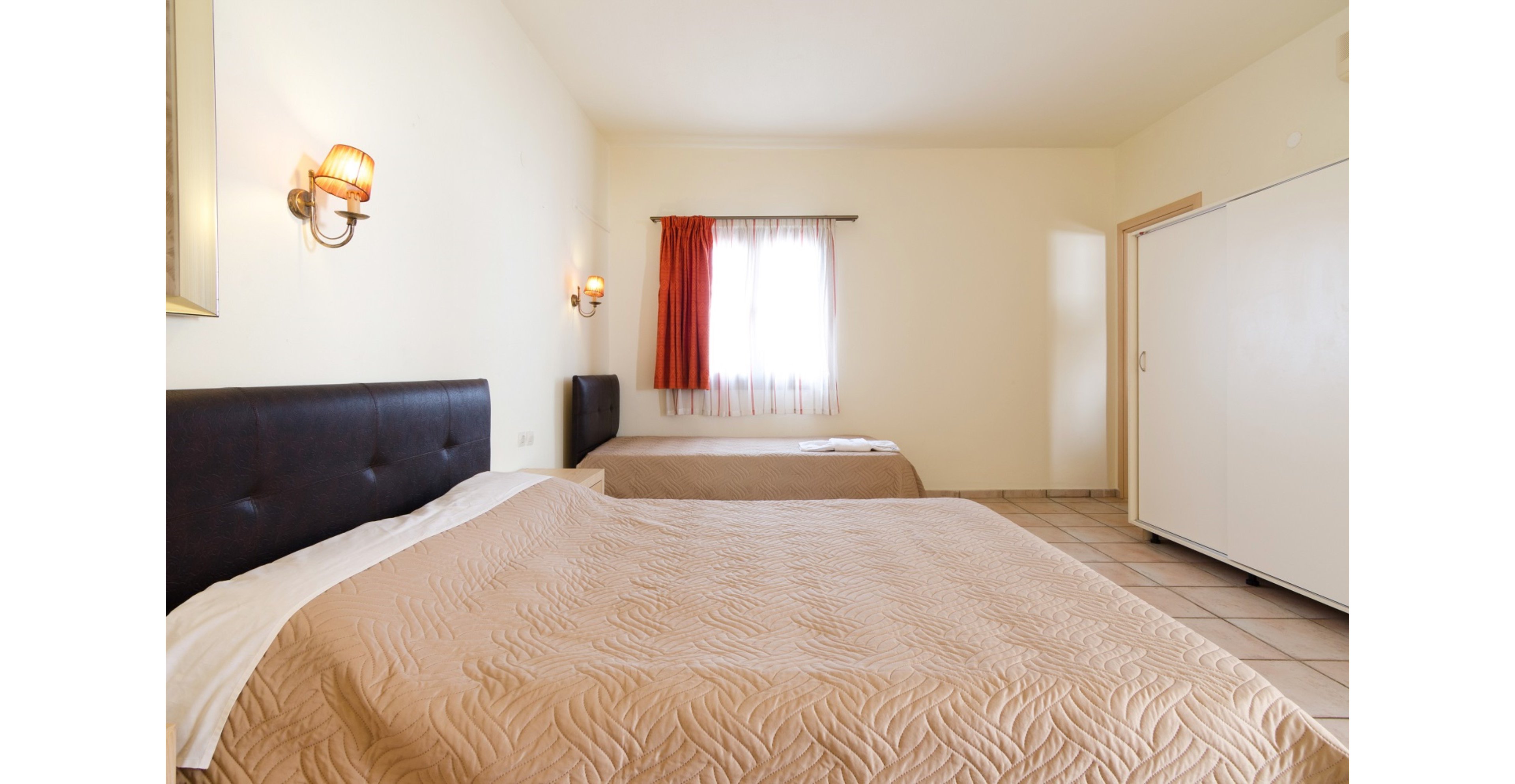 A simple hotel room features two double beds with beige quilted bedspreads and dark headboards. The space is brightly lit by natural light coming through a window with sheer white curtains and an orange drape. Wall-mounted lamps provide additional warm lighting on the pale cream walls. A white wardrobe and tiled floor complete the clean, minimalist interior.