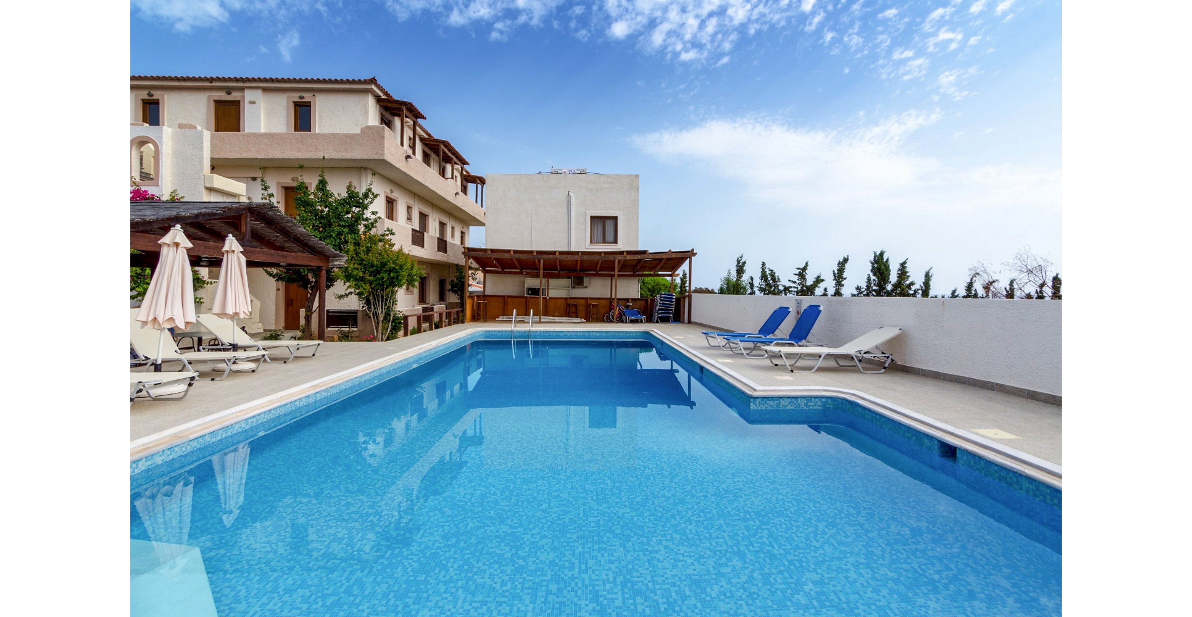 A clear blue swimming pool sits in the courtyard of a multi-story hotel or resort. Sun loungers and parasols line the tiled pool deck, with a shaded wooden pergola structure nearby. The building features balconies and warm-toned walls, framed by trees and a bright blue sky with scattered clouds. The scene suggests a relaxing vacation atmosphere in a quiet, well-kept outdoor space.