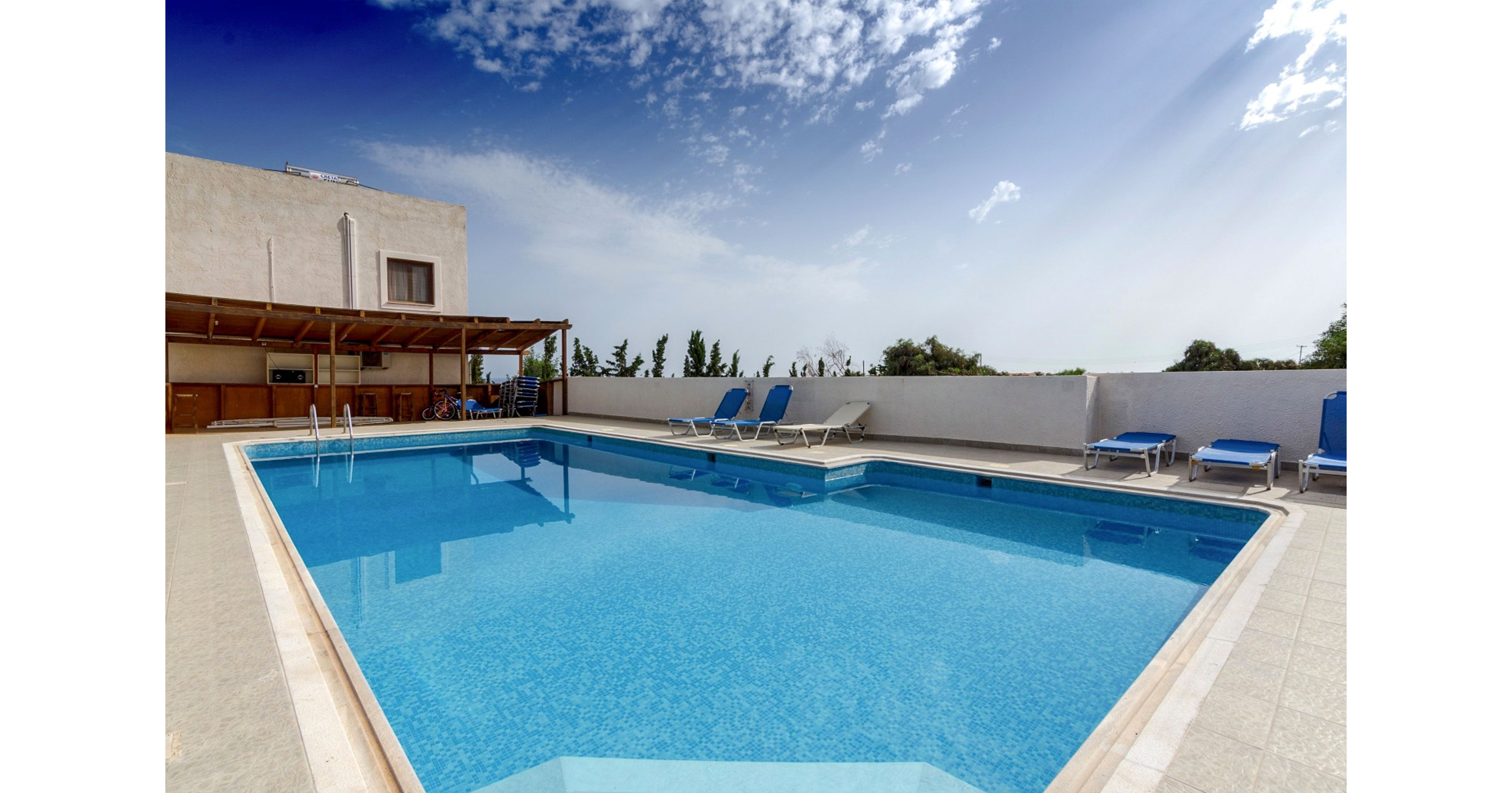 An outdoor swimming pool sits on a rooftop terrace under a bright blue sky. Several blue sun loungers line the poolside near a white wall, suggesting a relaxing leisure area. A shaded wooden pergola structure with seating and equipment is visible on one side of the terrace. The scene is calm and inviting, with clear water and soft sunlight creating a vacation atmosphere.